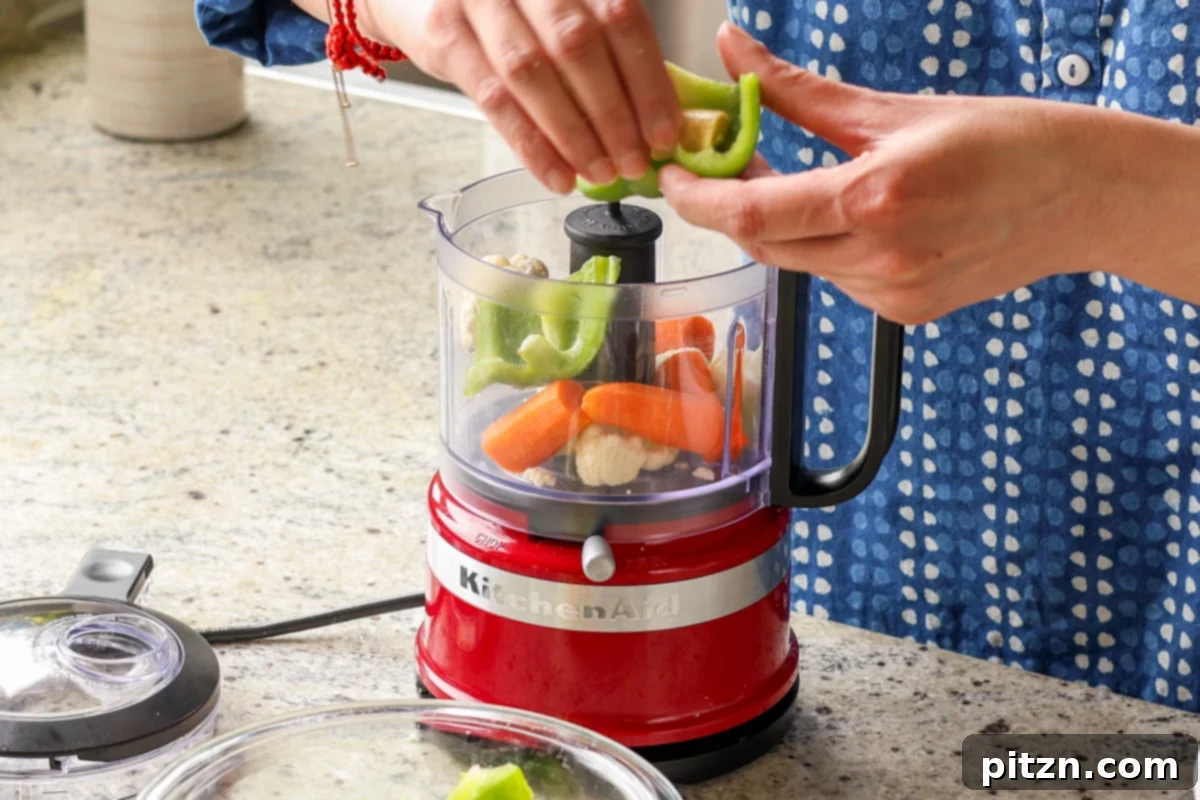 Adding finely chopped cauliflower, ginger, garlic, carrot, and bell peppers to a food processor for the lentil burger mixture.