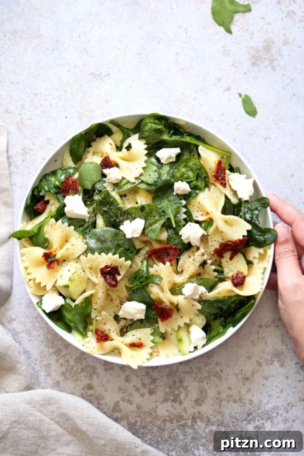 Tuscan Pasta Salad with Zesty Lemon Vinaigrette 2 Vibrant Tuscan Pasta Salad with sun-dried tomatoes, cucumber, spinach, and vegan feta, served in a white bowl on a concrete background.