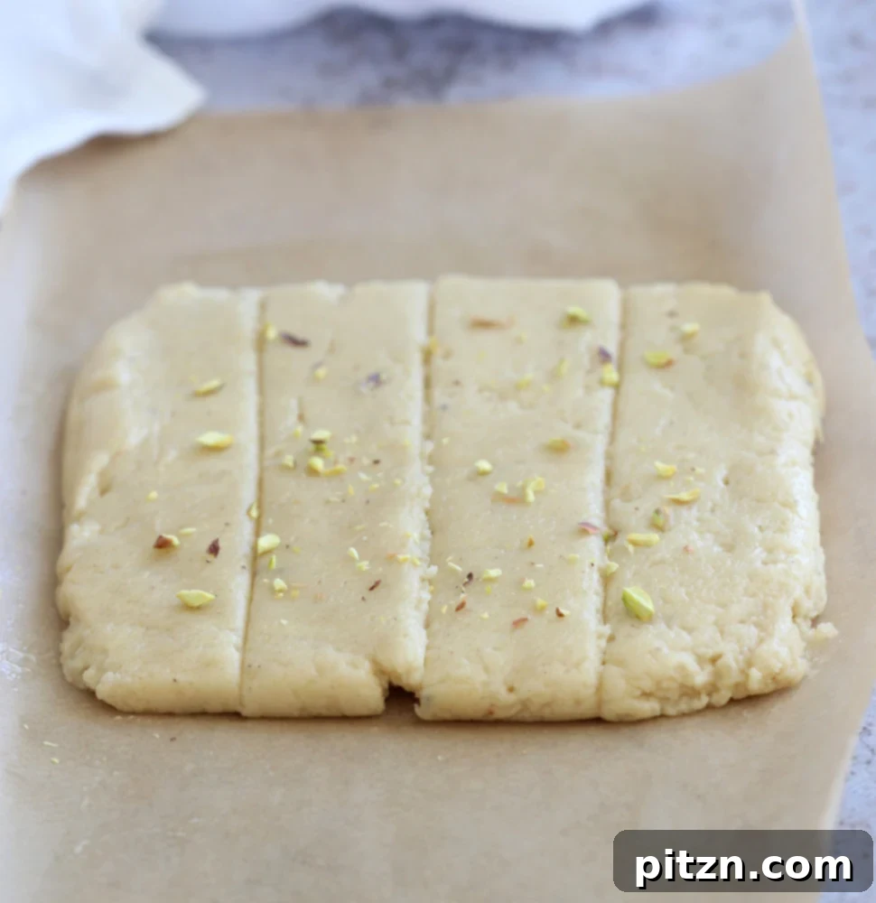 The shaped Vegan Malai Burfi mixture on parchment, pre-sliced and ready for baking, topped with pistachios.