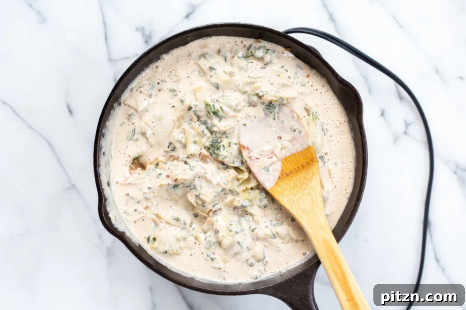 The smooth, creamy vegan sauce is being poured into the skillet, blending with the spinach and artichoke mixture to create the dip's rich texture.