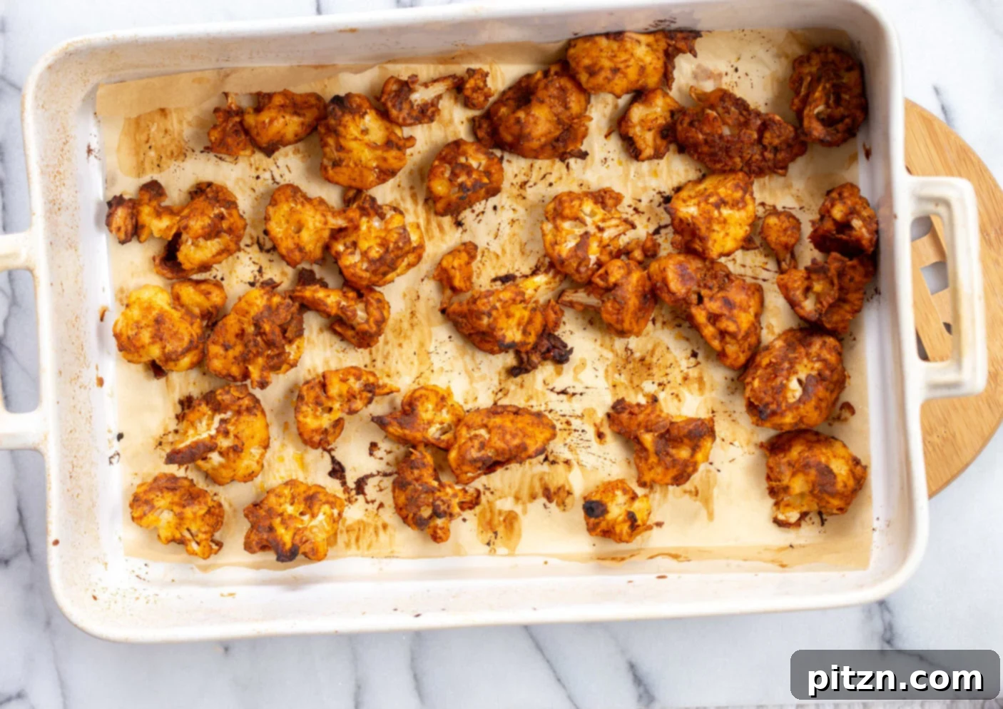 Our finished Vegan Nashville Hot Cauliflower Wings, glistening with spicy sauce on a white baking dish, garnished and ready to be served with refreshing dips and pickles.