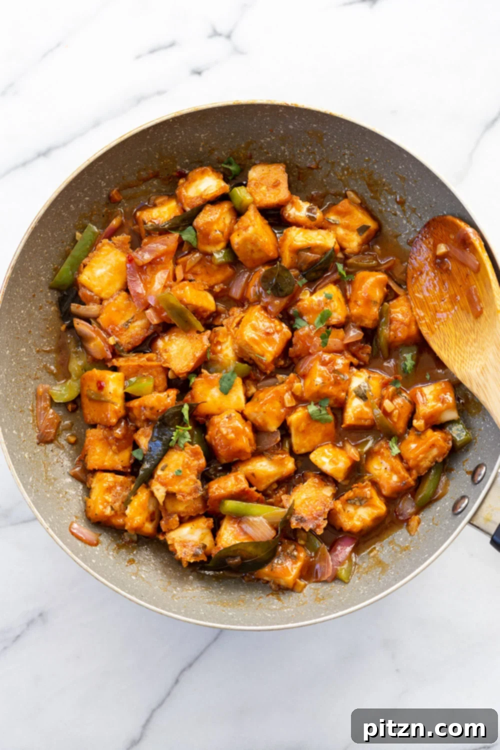 Tofu 65 in a grey skillet, showcasing perfectly browned, crispy tofu pieces glistening in the rich, thick sauce with visible chunks of onion and bell pepper. A delicious and wholesome vegan meal in progress.