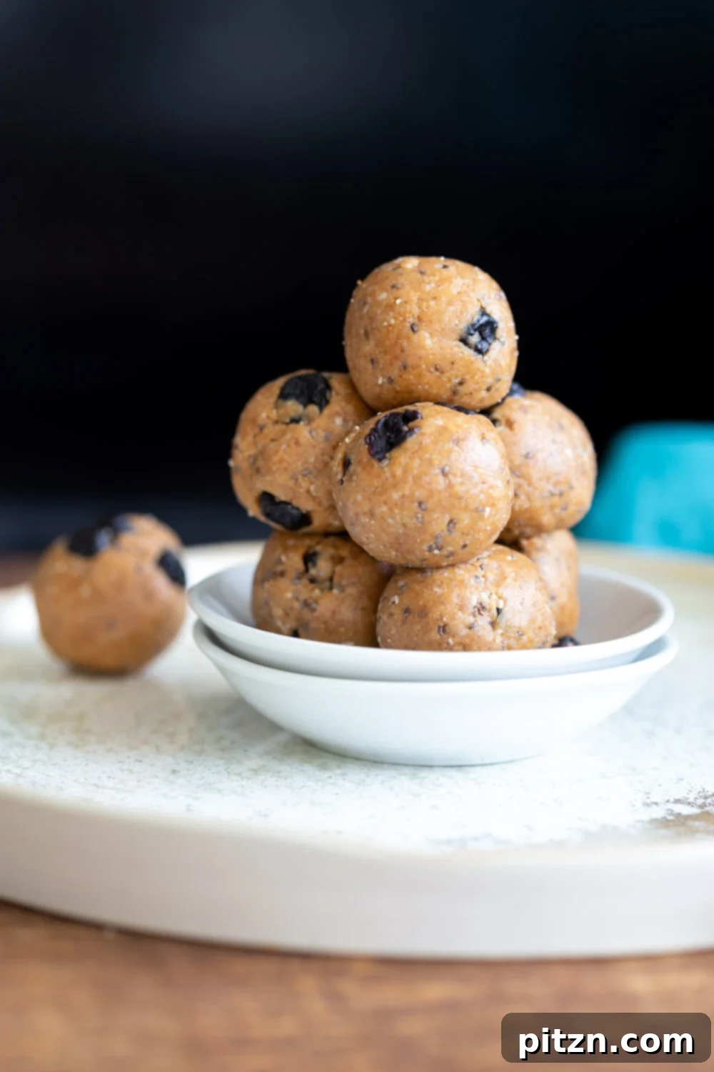 A stack of finished Vegan Blueberry Muffin Energy Bites on a white plate, set against a dark background, highlighting their delicious and ready-to-eat appearance.