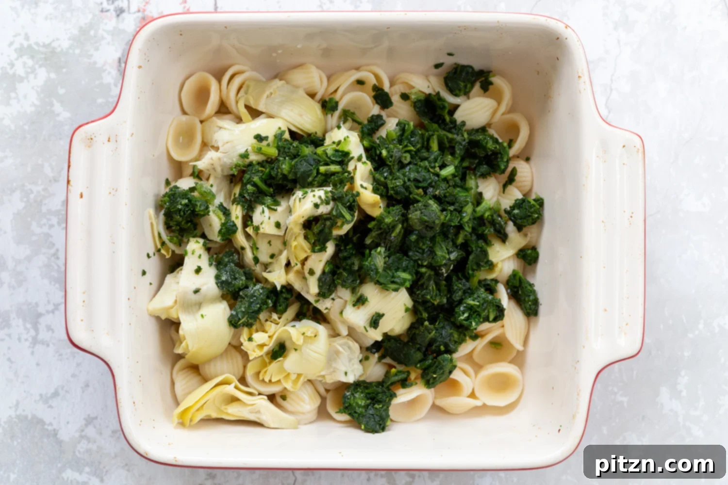 spinach pasta recipe overhead shot of a casserole dish with cooked shell pasta, spinach, and chopped artichokes