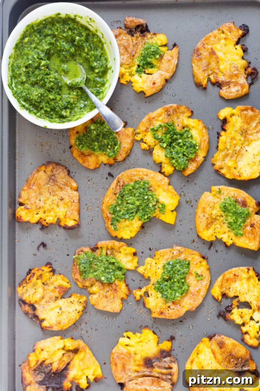 Crispy Vegan Smashed Potatoes with Chimichurri Sauce on a Grey Baking sheet, garnished with fresh herbs.