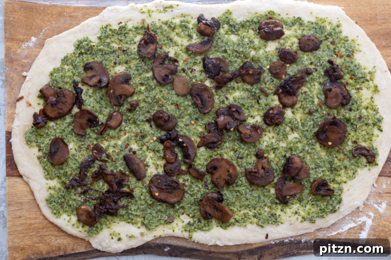 Adding Mushroom Filling Sautéed mushroom mixture being carefully sprinkled over the pesto-covered pizza dough, preparing it for rolling into savory pizza rolls.