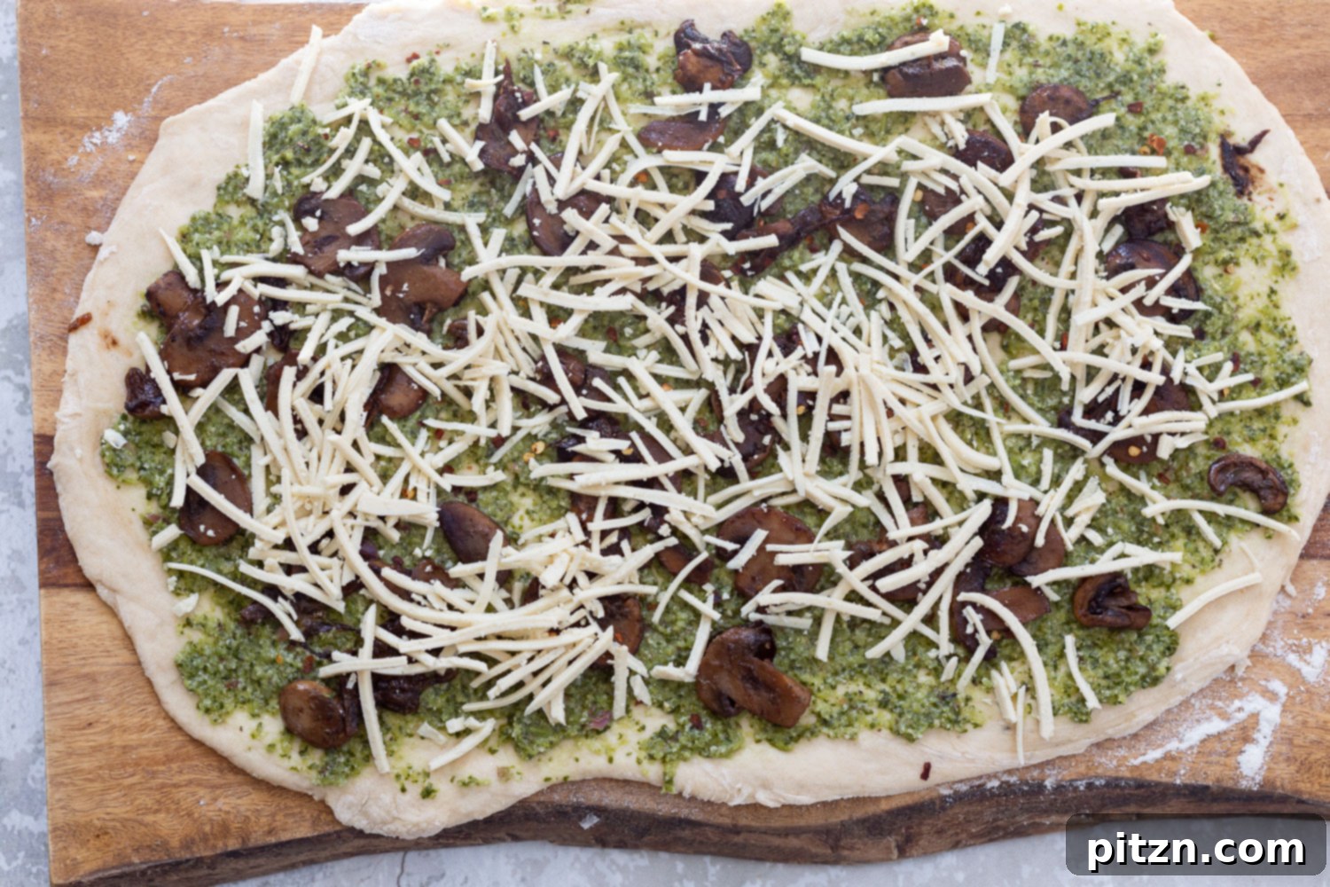 Adding Vegan Mozzarella Shredded vegan mozzarella cheese being generously sprinkled over pizza dough that's already layered with pesto and mushrooms, adding a cheesy finish before rolling.