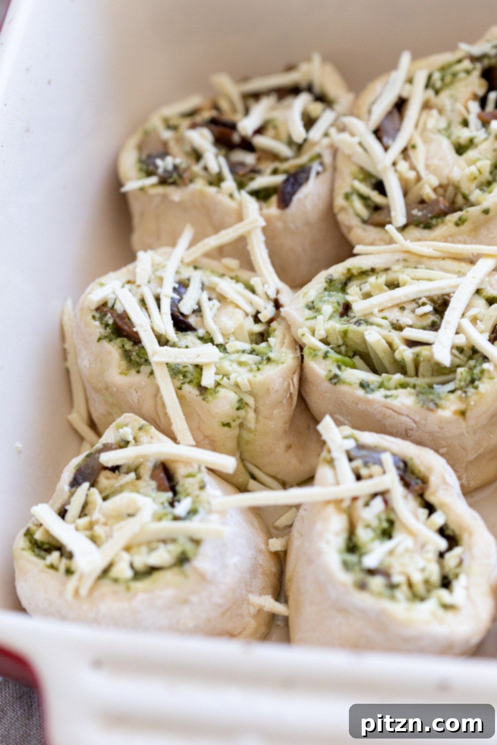 Unbaked Pizza Rolls Unbaked Pesto Pizza rolls arranged perfectly in a white ceramic baking dish, showing the vibrant green pesto and mushroom filling before they go into the oven.