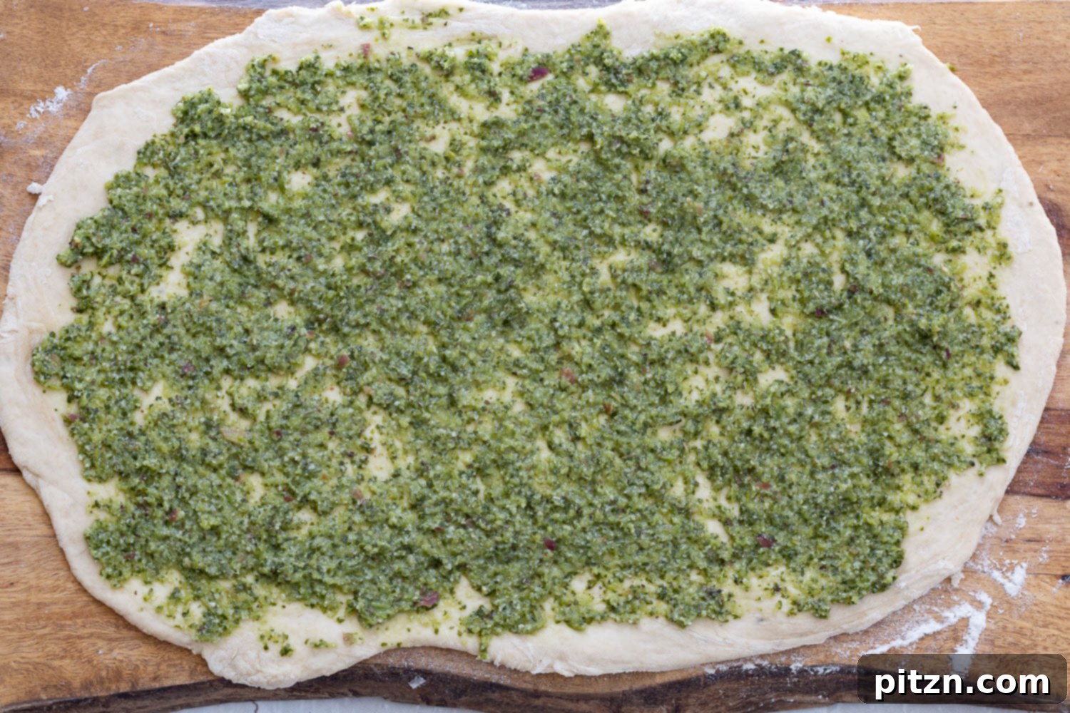 Spreading Vegan Pesto Vibrant green vegan pesto being smoothly spread across the surface of a rolled-out pizza dough rectangle, creating an even layer for the pizza rolls.