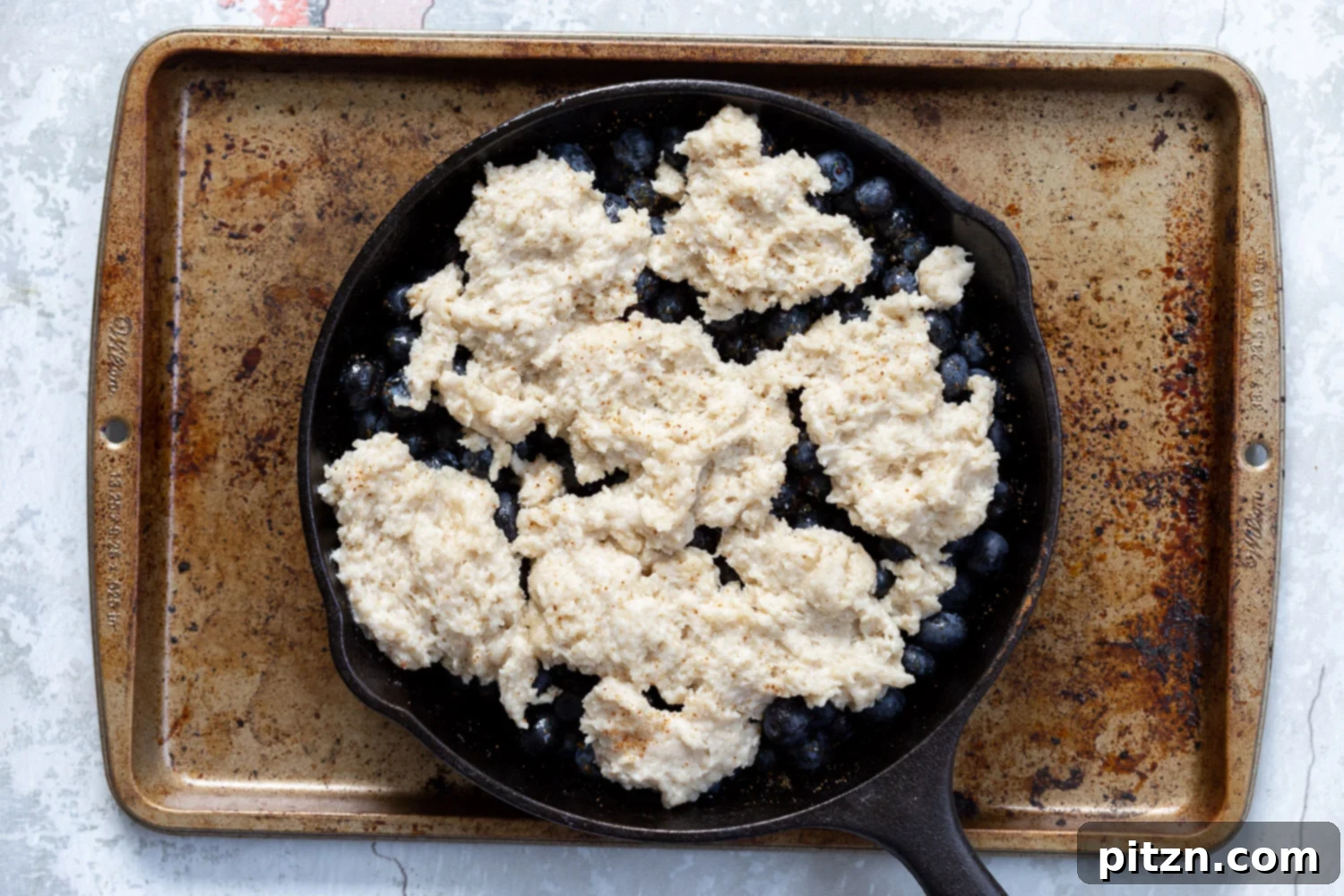 Assembled Vegan Blueberry Cobbler in a cast iron skillet, placed on a baking sheet, ready to be put into the oven.