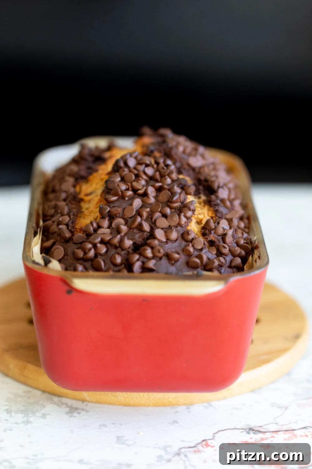 A whole loaf of Vegan Marbled Chocolate Banana Bread, generously topped with chocolate chips, baked to golden perfection in a vibrant red ceramic loaf pan.