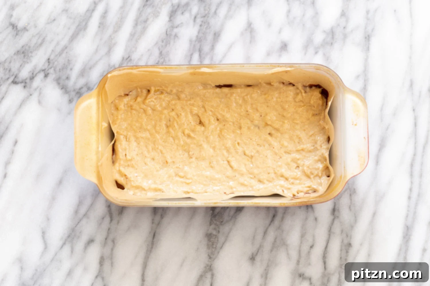 A scoop of the lighter vanilla banana bread batter being carefully placed on top of the darker chocolate batter in a loaf pan, demonstrating the layering technique for marbling.