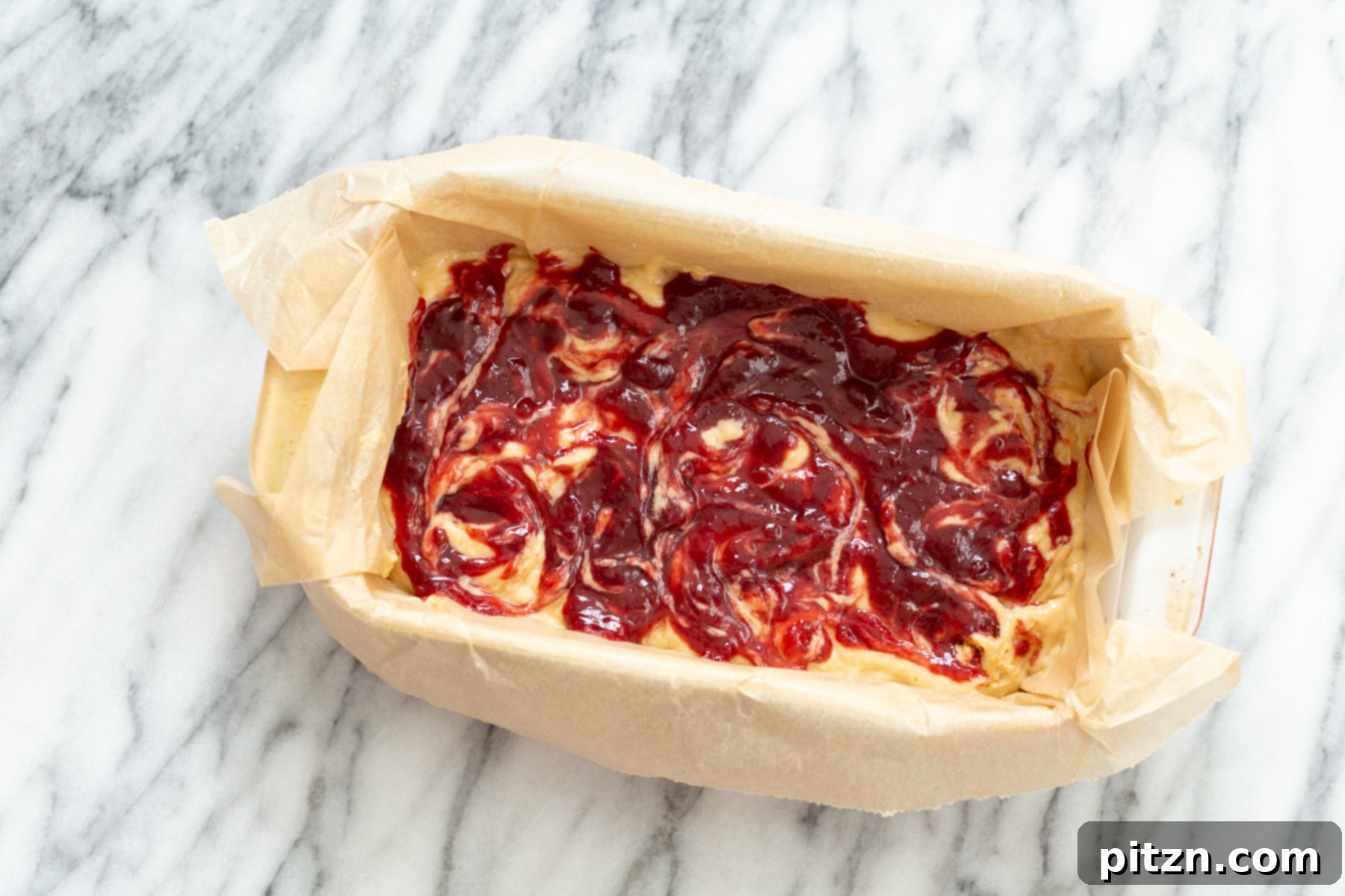 Vibrant raspberry jam being artistically swirled into rich peanut butter cake batter in a mixing bowl, creating beautiful marbled patterns.