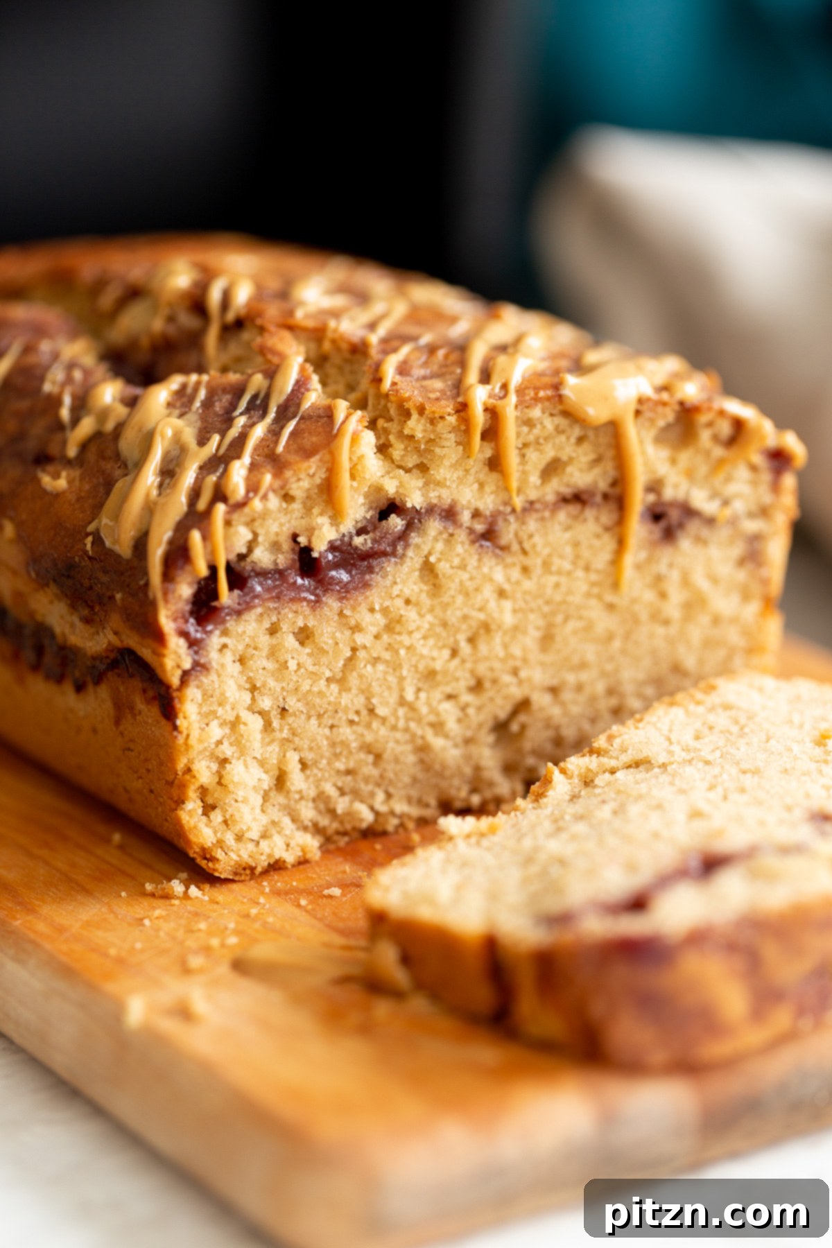 A close-up view of a sliced vegan PB&J loaf cake, beautifully drizzled with extra peanut butter on a wooden cutting board, highlighting its moist texture and jam swirl.