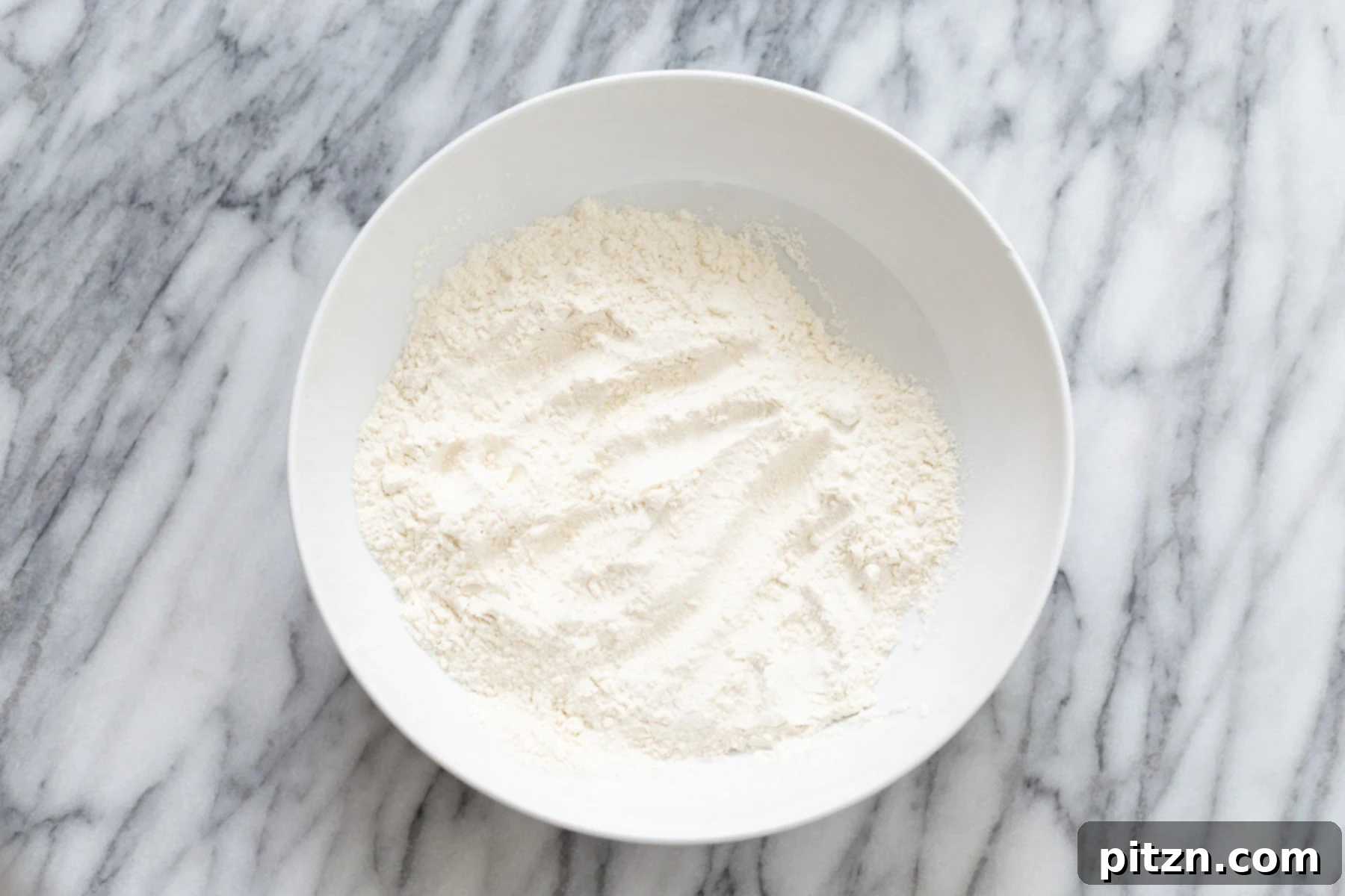 A neatly arranged white bowl filled with dry cake mix, including flours, salt, baking powder, and baking soda, ready for combination.