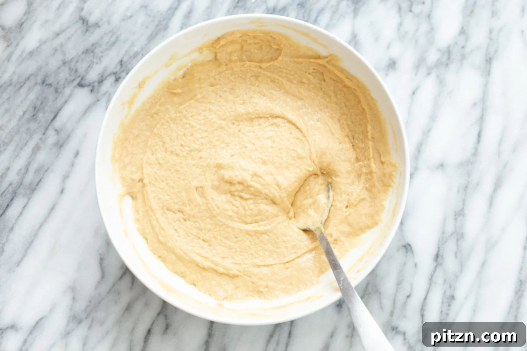 Creamy peanut butter cake batter being gently mixed with a metal spoon in a white ceramic bowl, showcasing its smooth texture.