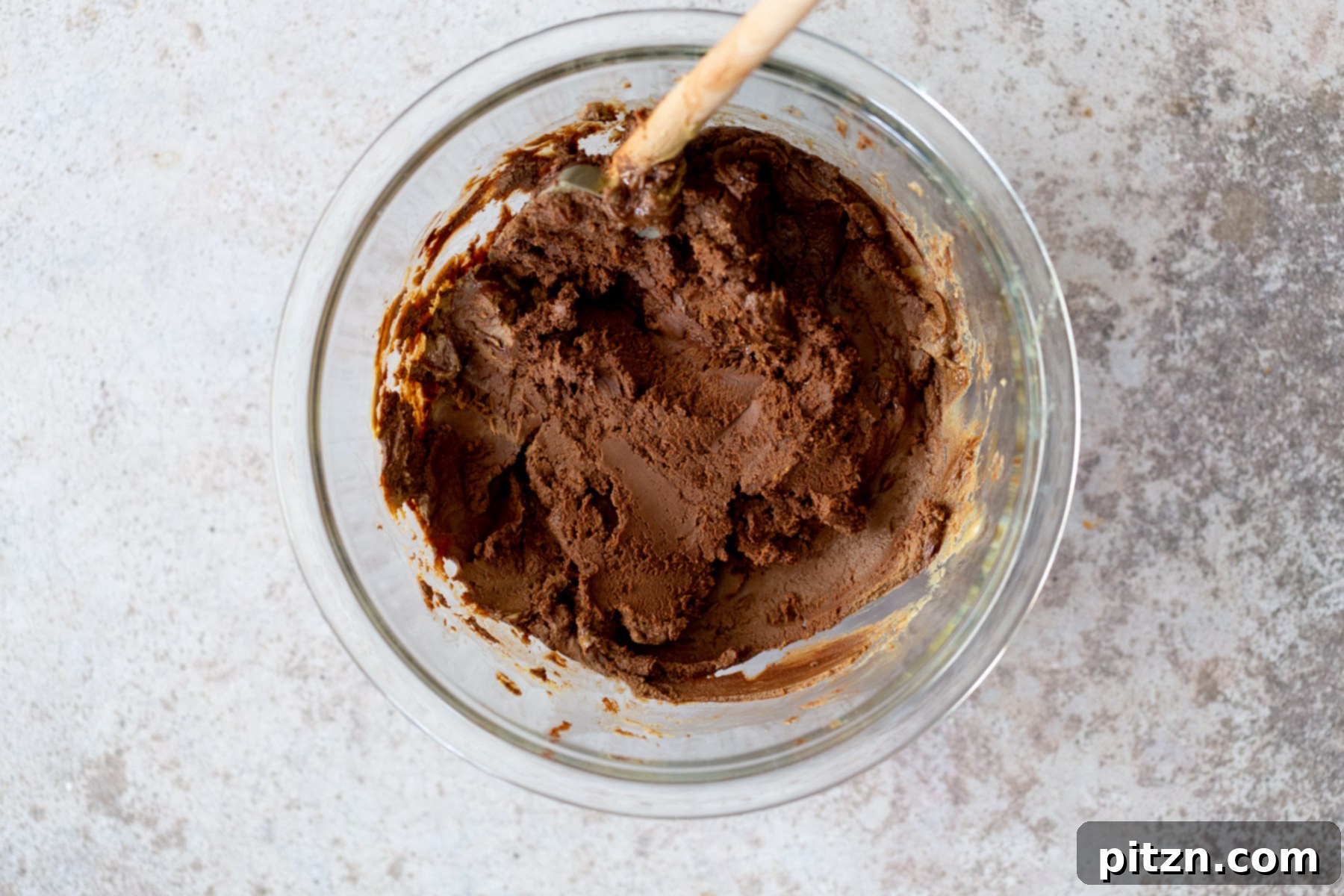 Mixing vegan fudge batter in a clear glass bowl over a double boiler, showing the chocolate and nut butter melting together to create a smooth, glossy mixture.