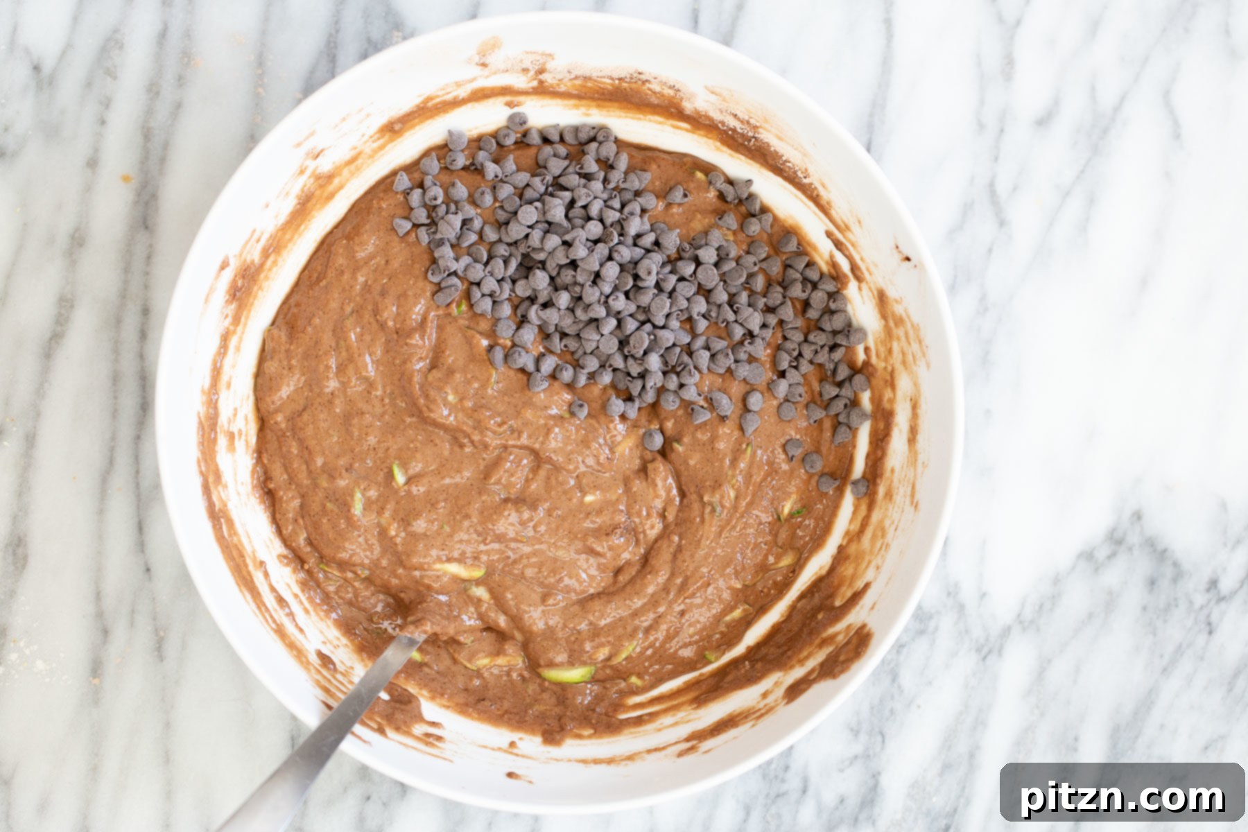 Overhead shot of a white bowl filled with vegan zucchini bread batter, generously sprinkled with dairy-free chocolate chips before baking.