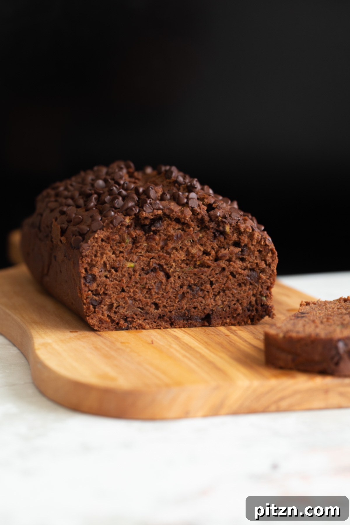 A side view of a moist, rich vegan chocolate zucchini bread loaf with one slice cut, revealing its soft, dark crumb and chocolate chips.
