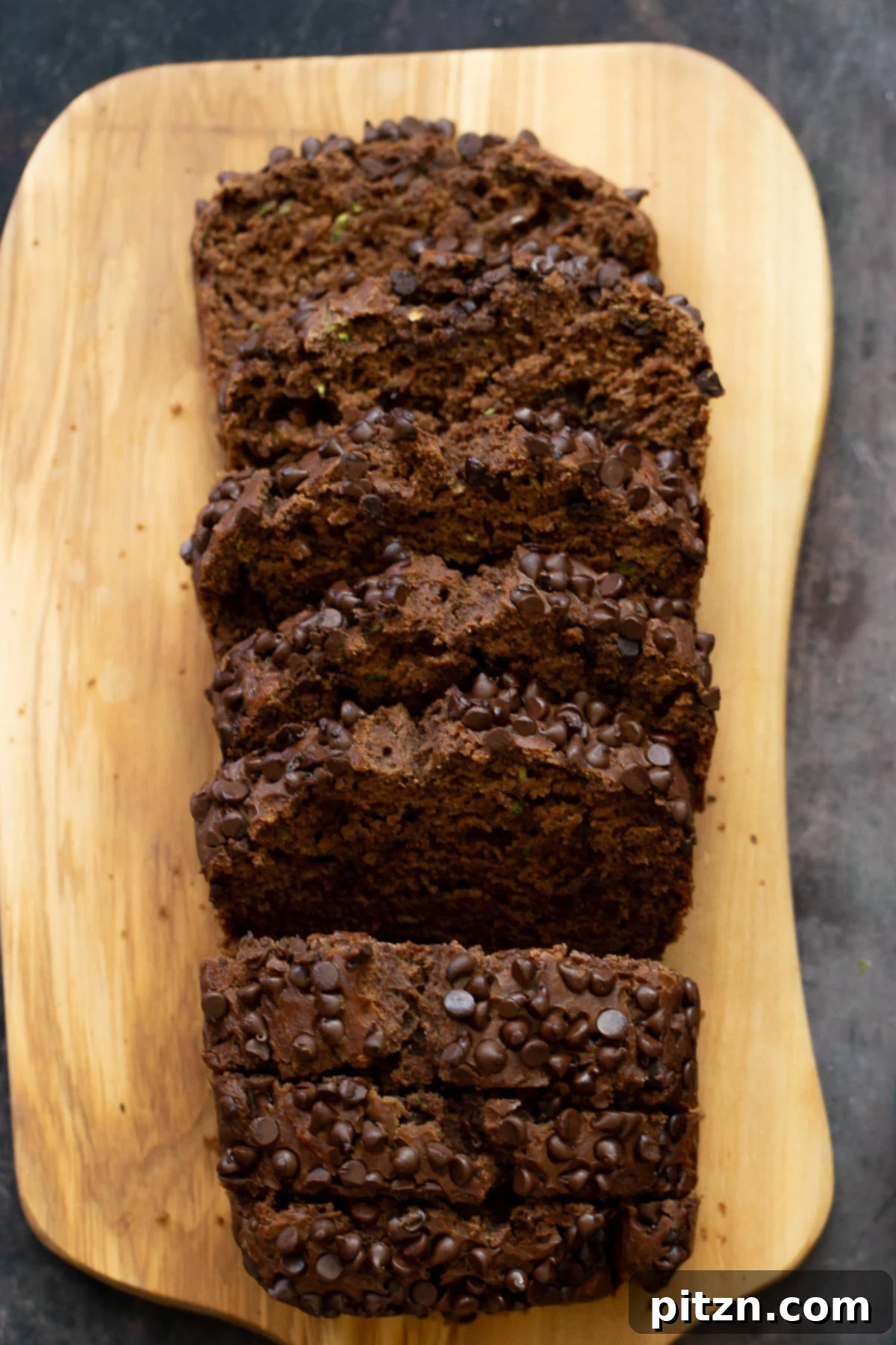 Overhead shot of a perfectly sliced loaf of vegan chocolate zucchini bread on a wooden chopping board, revealing its moist interior and scattered chocolate chips.
