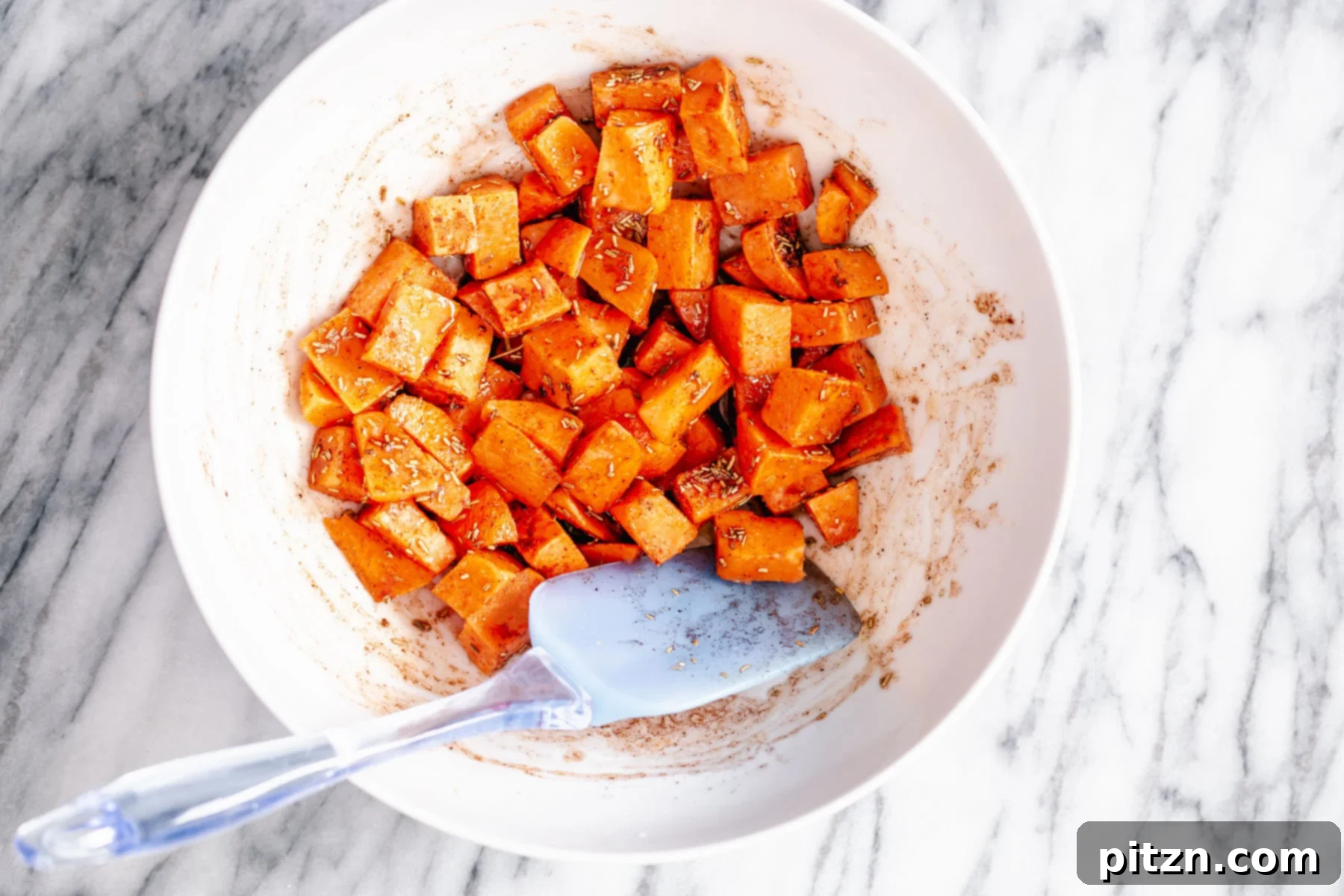 Tossing Sweet Potatoes Sweet potato cubes are being tossed in a white bowl, coated evenly with spices and maple syrup, ready for roasting.