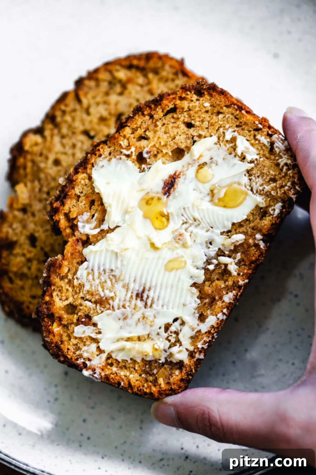 A hand reaching for a slice of warm, buttered vegan gingerbread banana bread, highlighting its inviting texture.