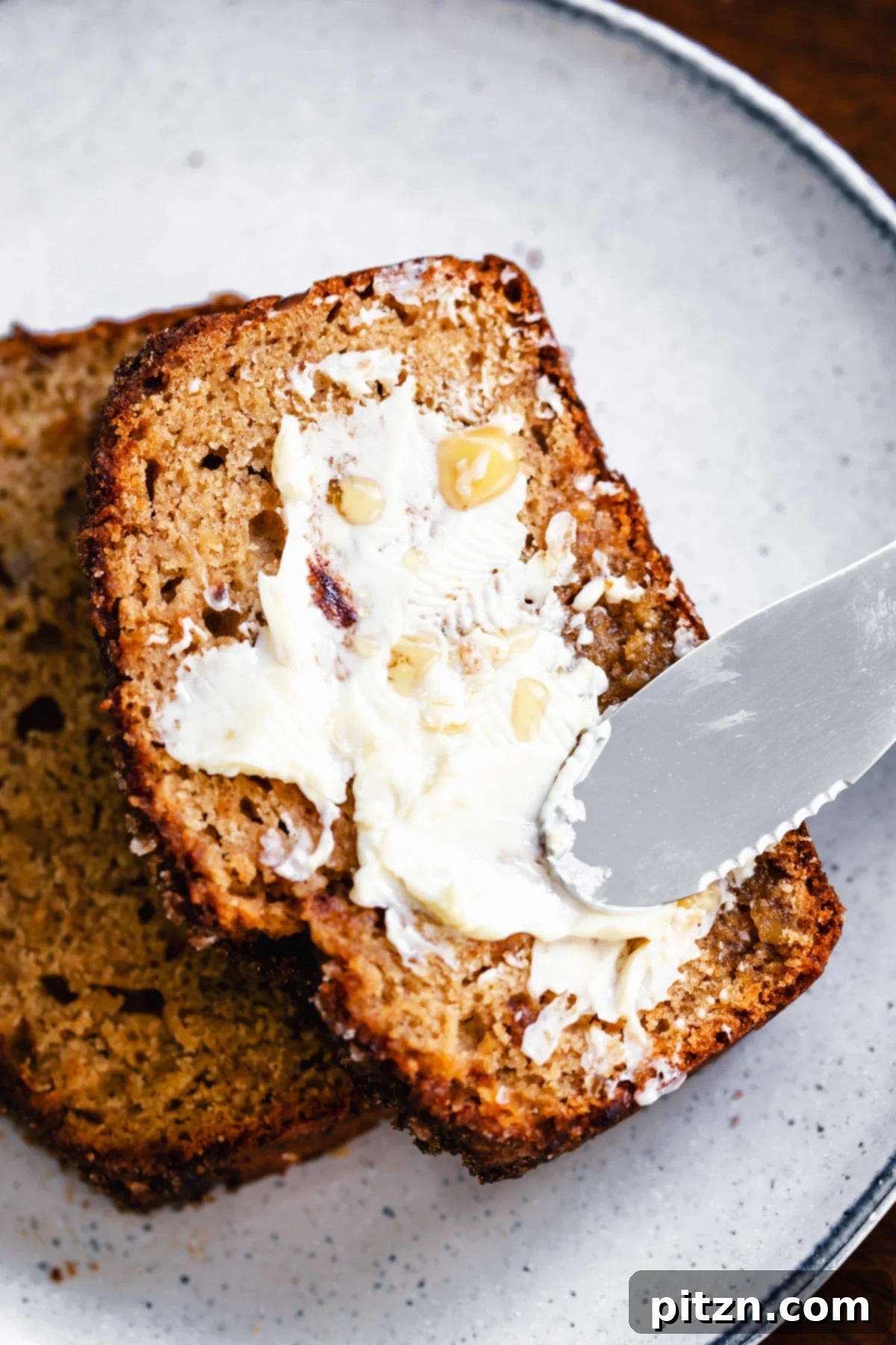 A close-up shot of a slice of vegan gingerbread banana bread being delicately buttered, showing its rich, spiced interior.