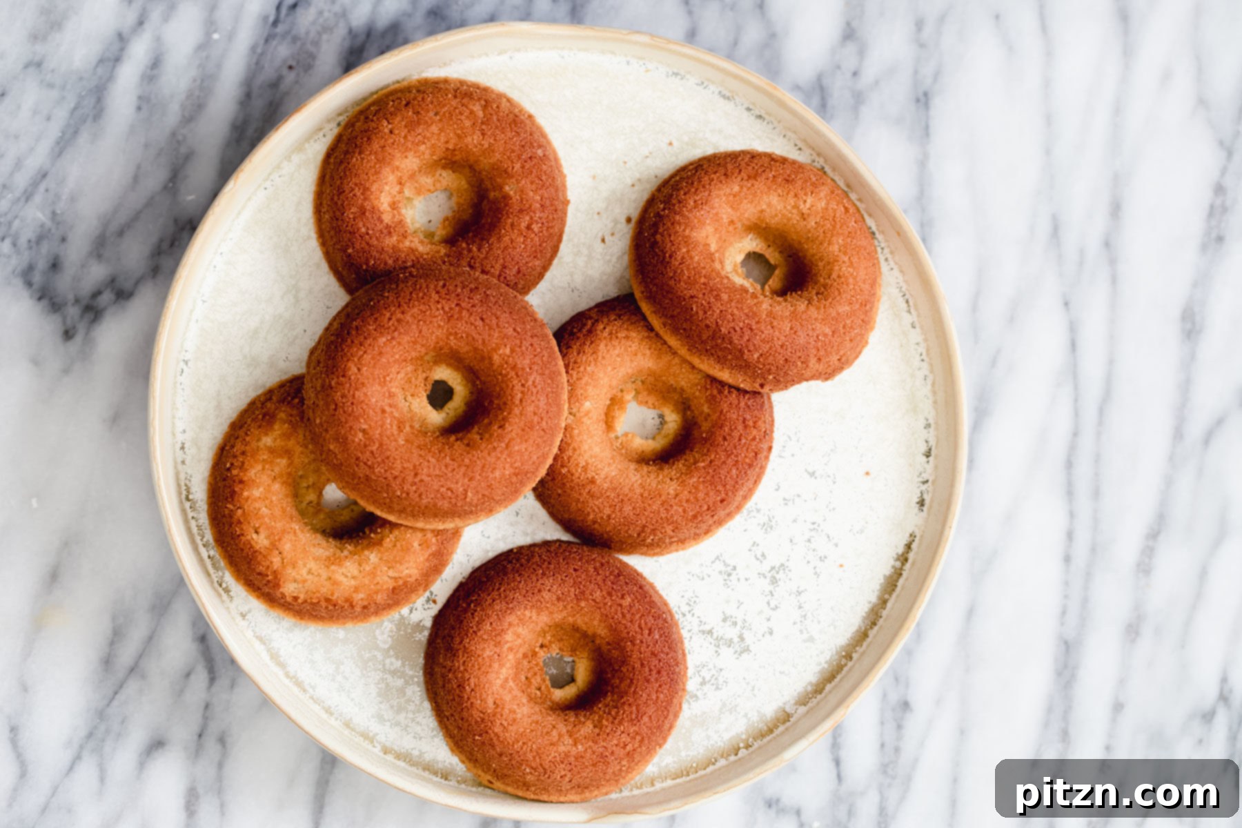 Gluten-Free Donuts on Marble Table An overhead shot of a pristine white plate adorned with an array of freshly baked gluten-free donuts, glistening with a cinnamon sugar coating, all elegantly placed on a marble kitchen table.
