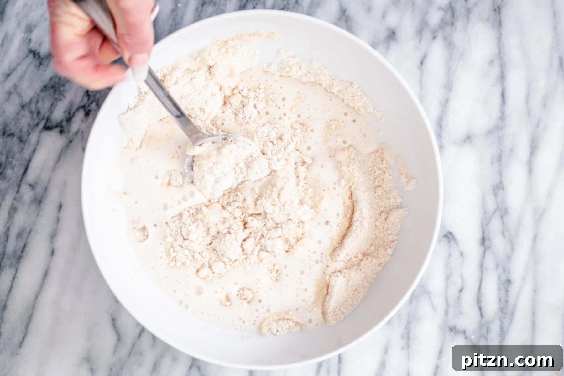 Mixing Wet into Dry Ingredients for Donuts Wet ingredients are being gently stirred into the dry donut mixture in a white bowl, showing the gradual incorporation to form a smooth, lump-free batter for gluten-free vanilla donuts.