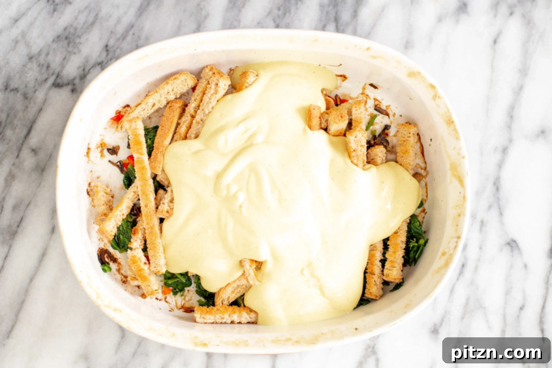 Vegan Eggs Benedict Breakfast Casserole 2 The smooth, golden vegan egg sauce made from tofu being generously poured over the layered ingredients in the breakfast casserole dish.