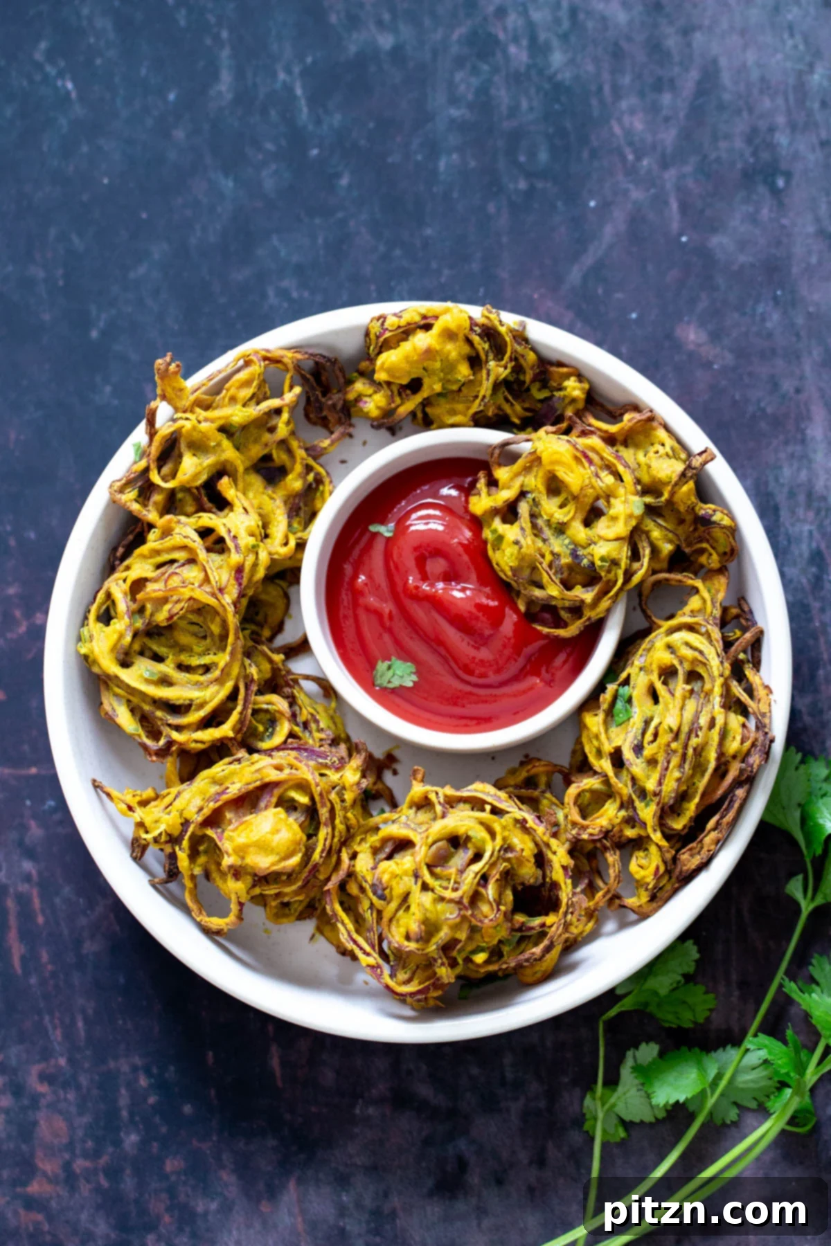 A vibrant overhead shot of golden-brown baked onion pakora served on a platter, accompanied by a small bowl of classic ketchup for dipping. Each pakora boasts a perfectly crispy exterior and tender, flavorful onion within.