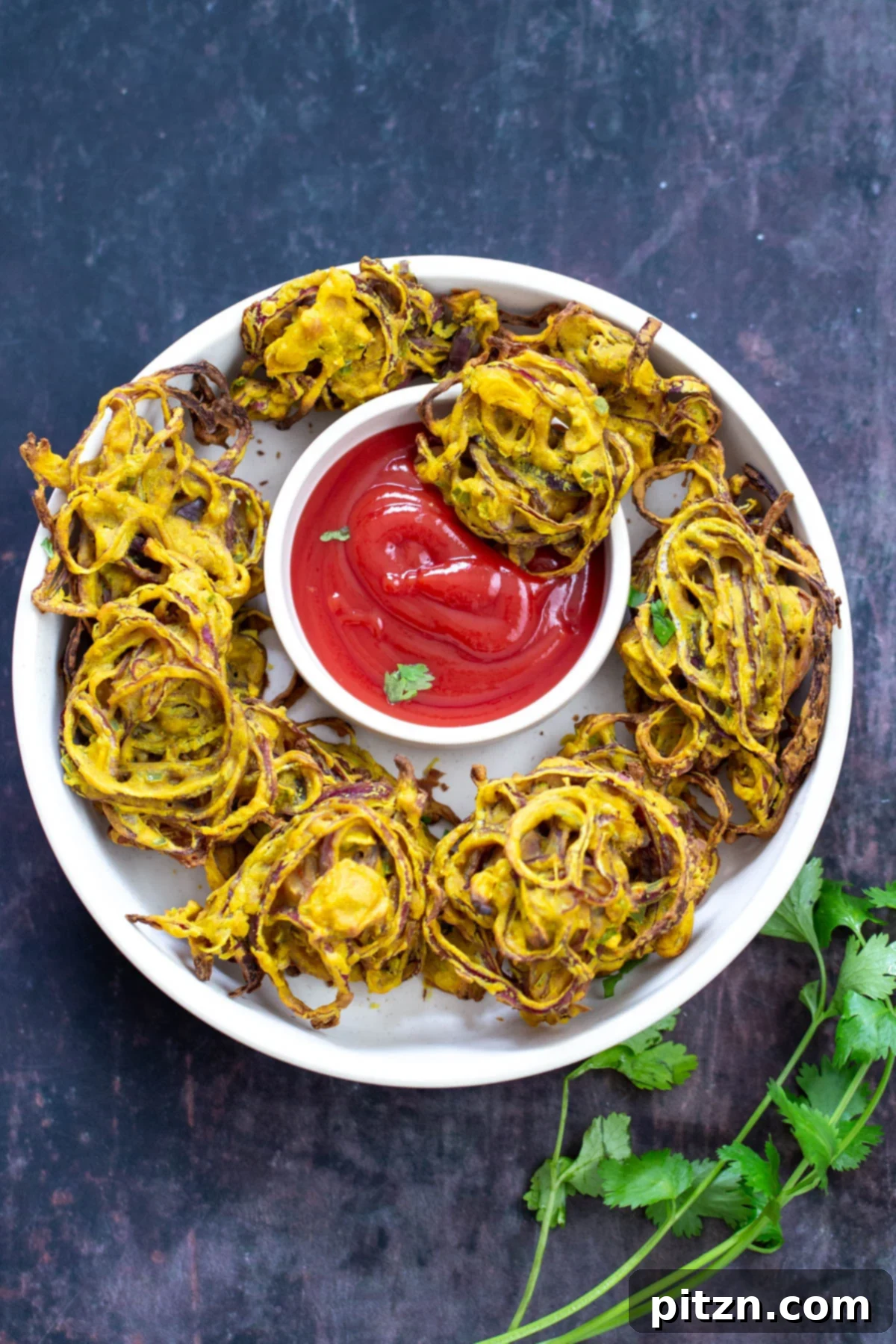 A close-up of beautifully arranged baked vegan onion bhajis on a pristine white platter, with a vibrant red dish of ketchup nestled in the center as a dipping sauce. The golden pakoras are enticingly crisp.