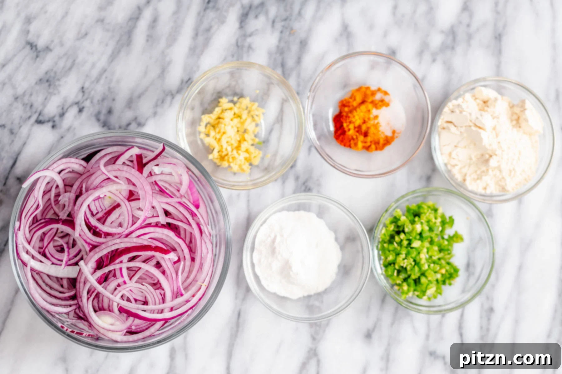 A flat lay photograph showcasing all the fresh ingredients meticulously arranged and ready for making baked pakora. Thinly sliced red onions, a variety of vibrant spices, chickpea flour, and rice flour are visible, highlighting the simplicity and natural goodness of the recipe.