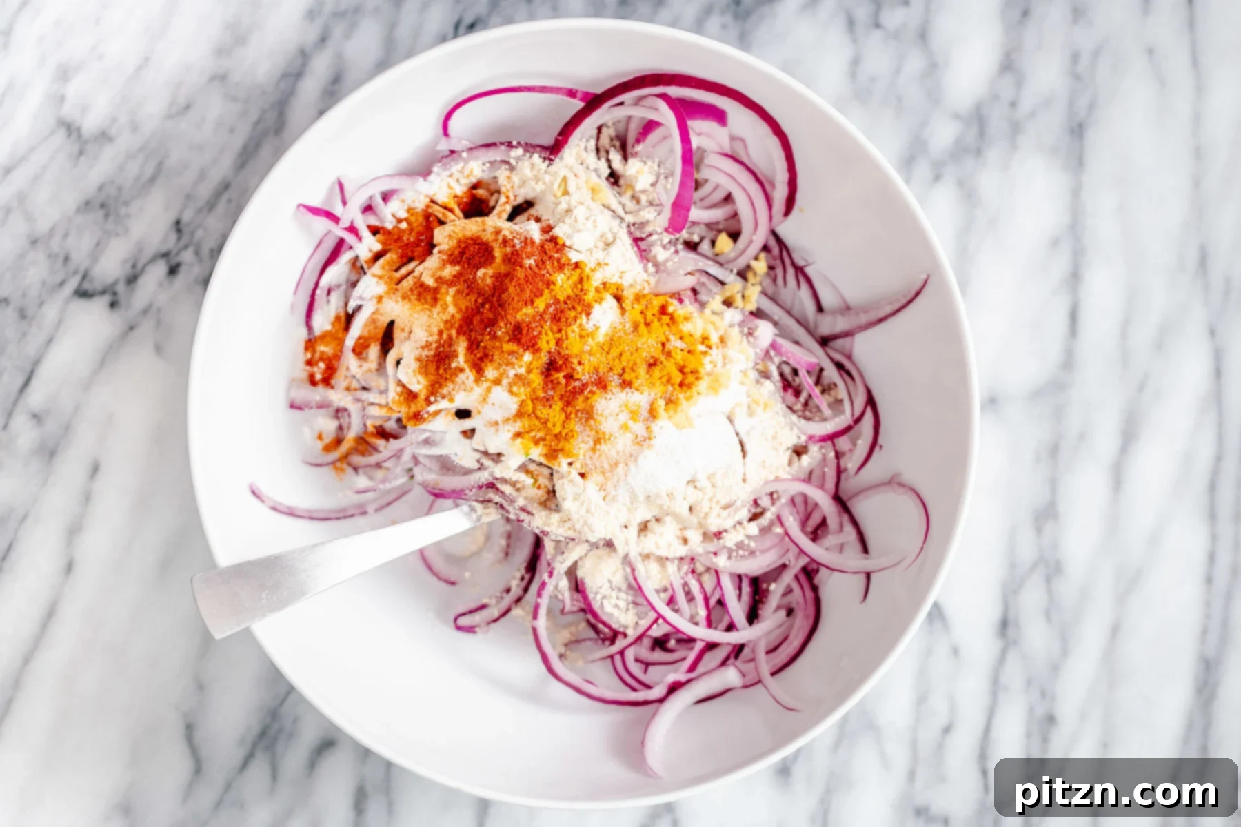 A close-up shot of thinly sliced red onion rings being gently mixed with chickpea and rice flour in a large bowl. The flours lightly coat the onion, preparing for the next stage of making crispy onion bhaji.
