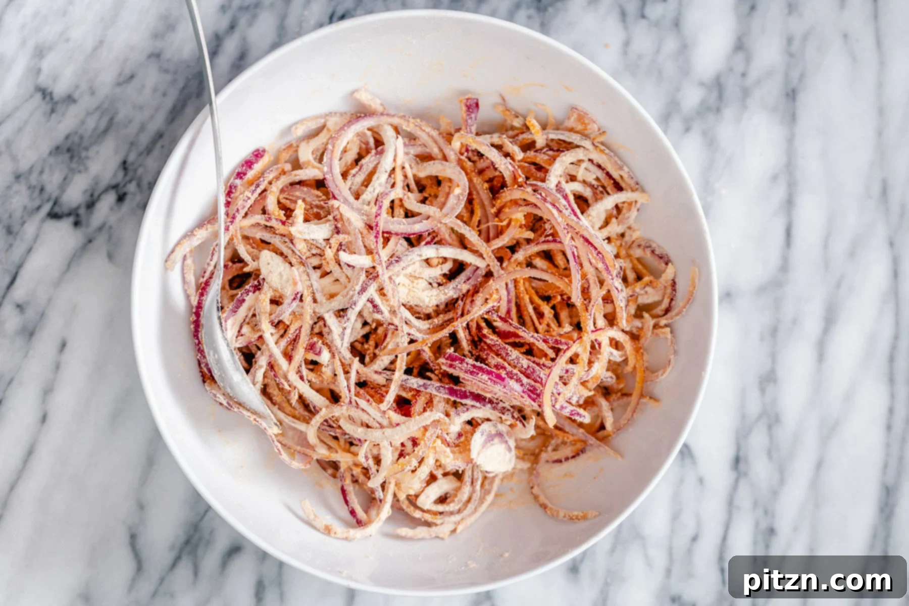 A bowl of thinly sliced onions coated in a vibrant mix of chickpea flour and various Indian spices, being expertly combined by hand. The mixture is prepped for the addition of oil and water to form the pakora batter.