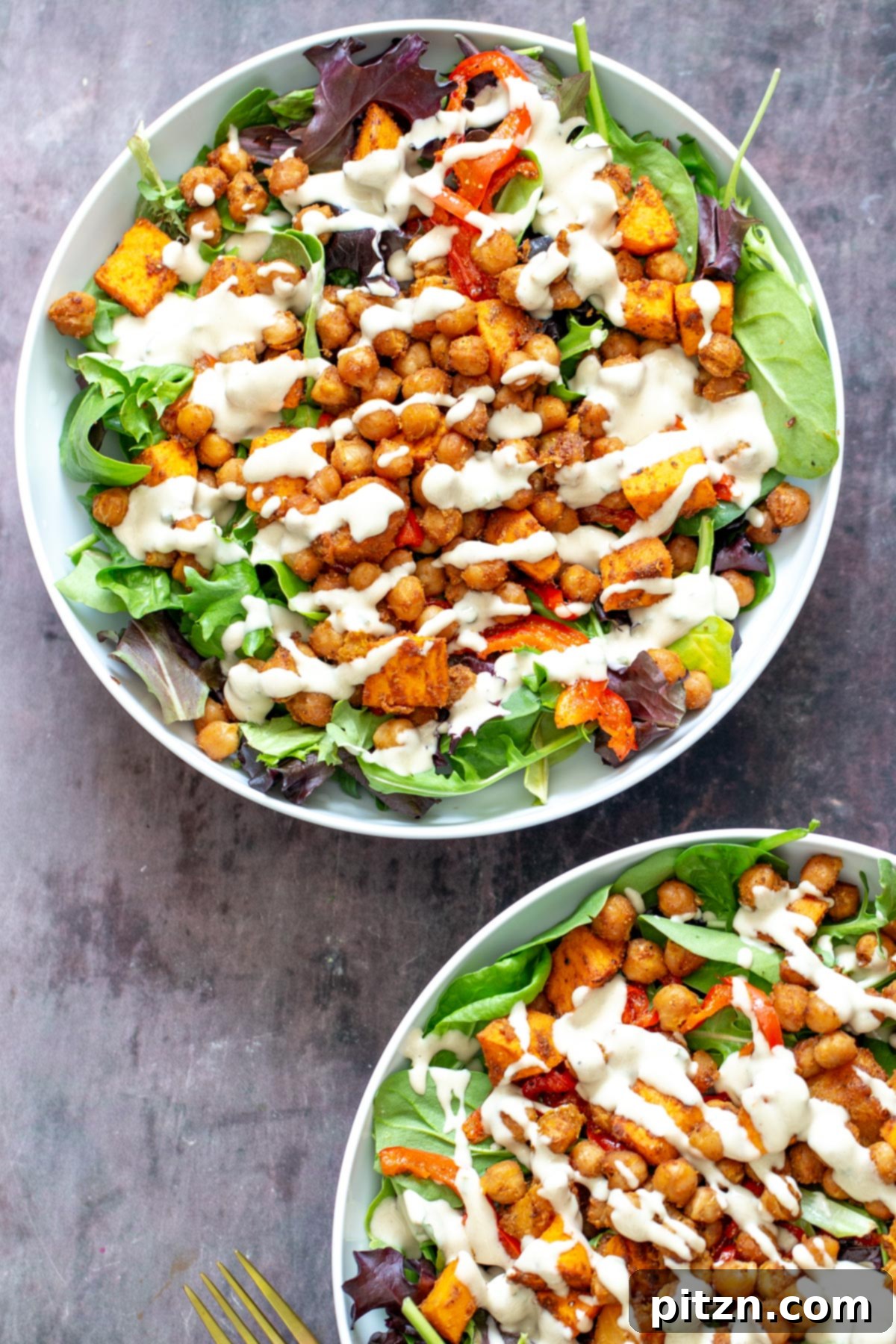 Two vibrant salad bowls featuring a spring mix base, topped with perfectly roasted chickpeas, bell peppers, and sweet potatoes, generously drizzled with tahini dressing.