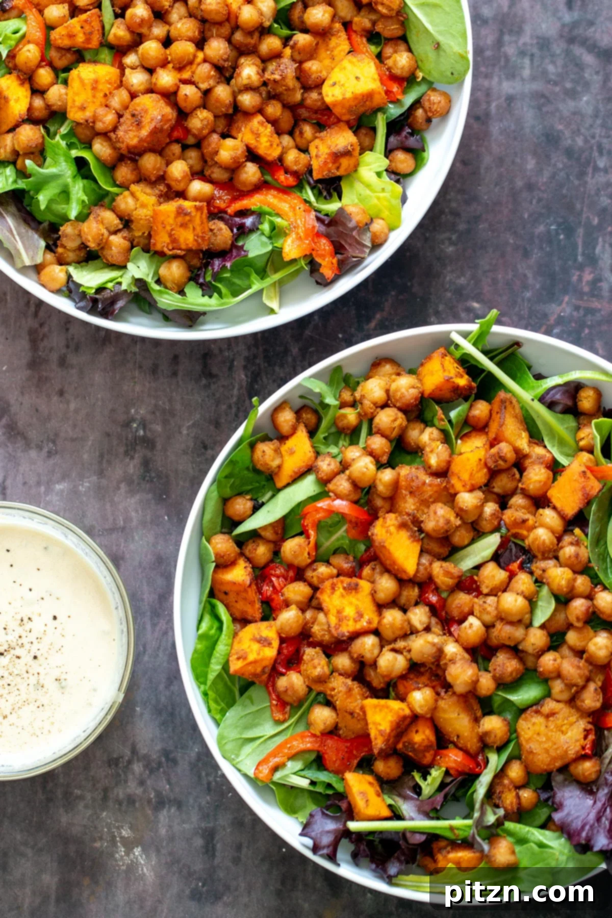 Two salad bowls featuring baked chickpeas, roasted bell pepper, and sweet potatoes, accompanied by a small dish of creamy tahini dressing, ready to be served.