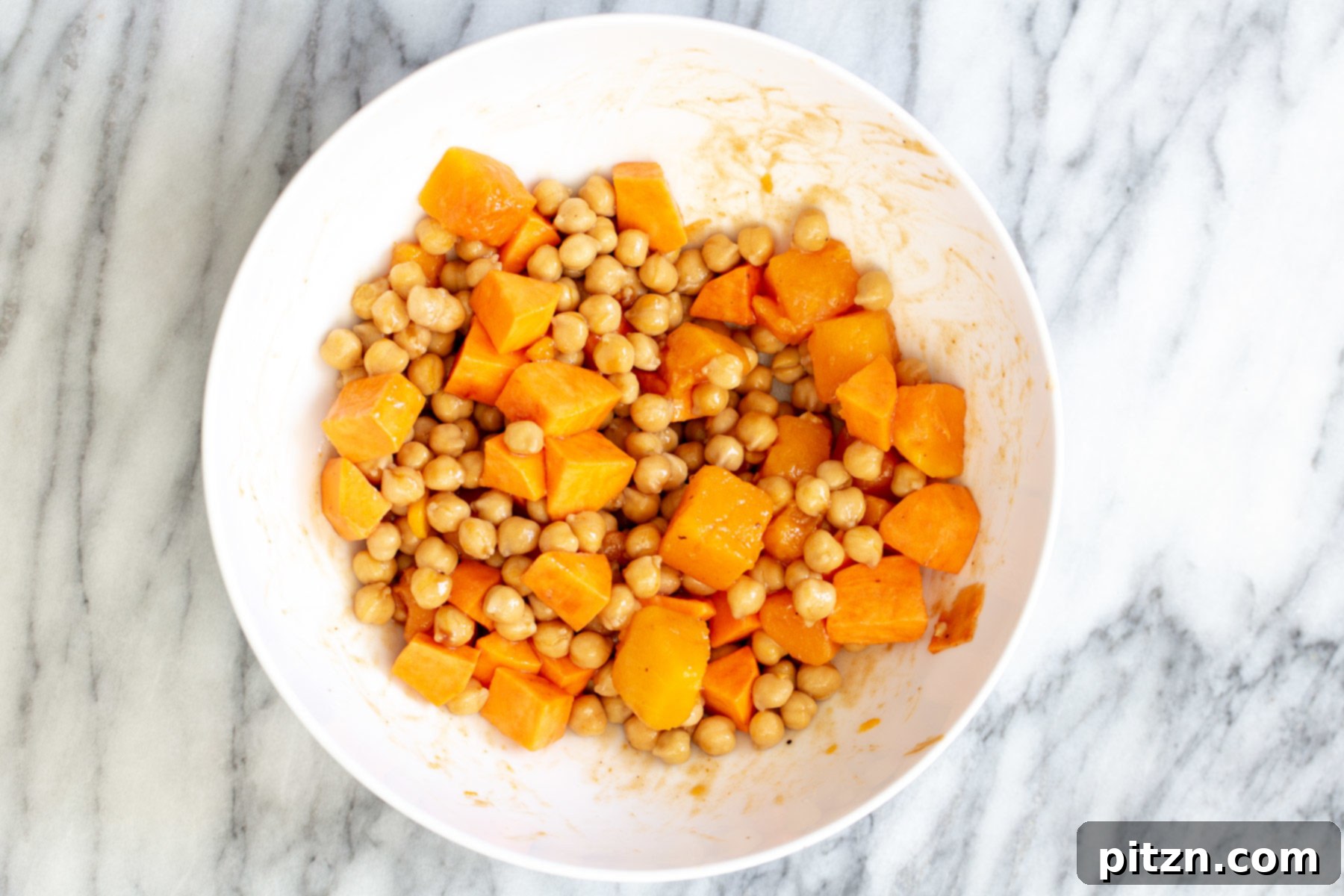 Sweet potatoes and chickpeas being mixed in a bowl with BBQ sauce and oil, ensuring an even coating before adding spices.
