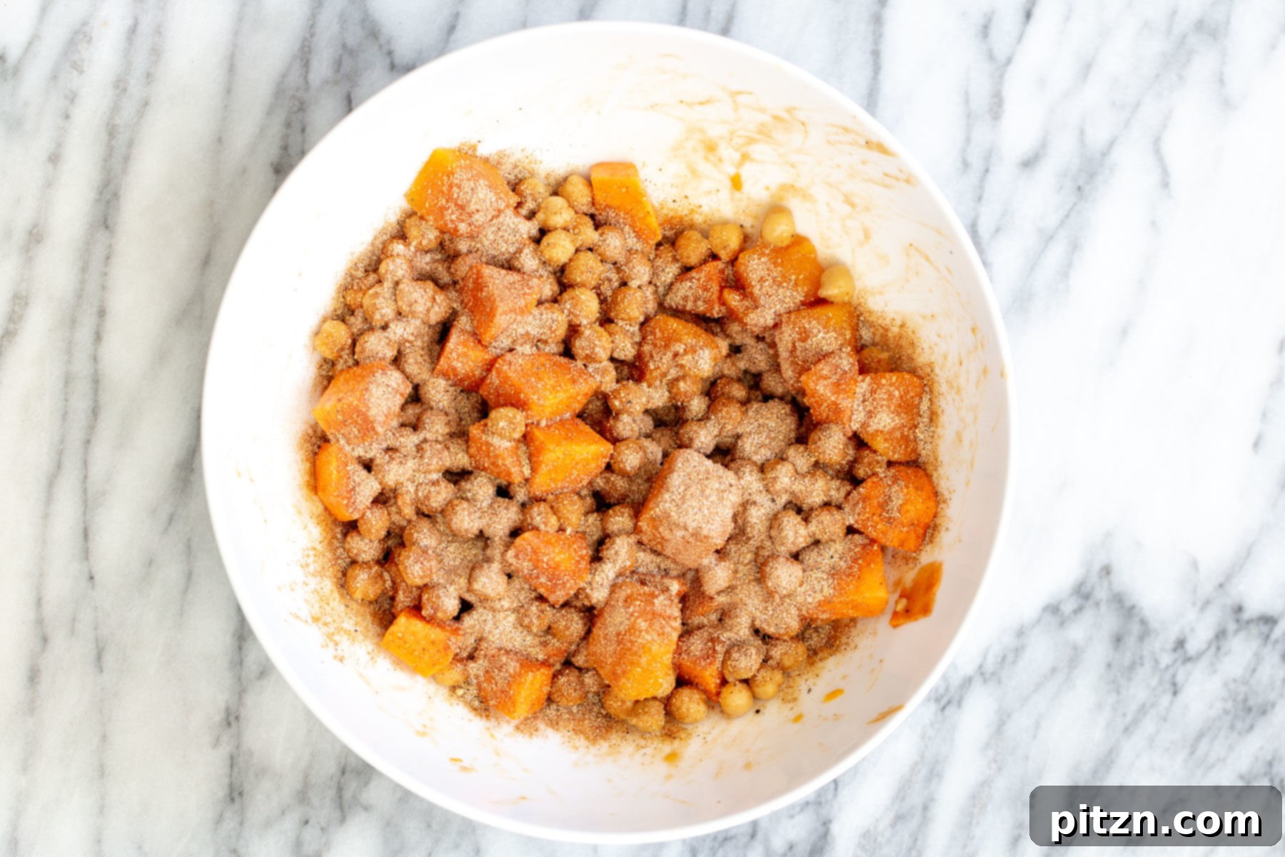 A vibrant spice mix being sprinkled generously on top of sweet potatoes and chickpeas in a bowl, ready for mixing.