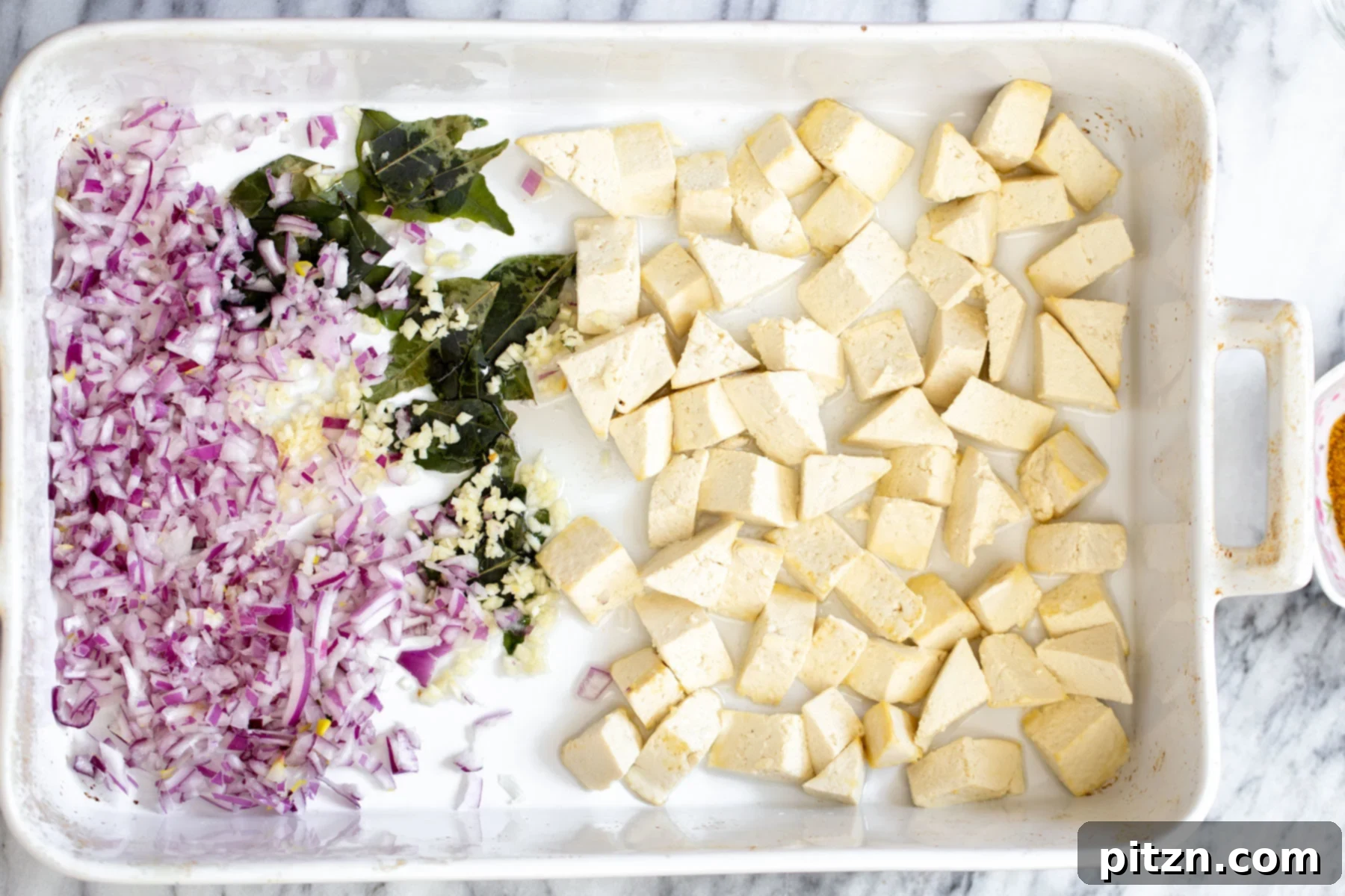 A casserole dish with perfectly cubed tofu, finely chopped onions, and fragrant curry leaves, ready for seasoning.