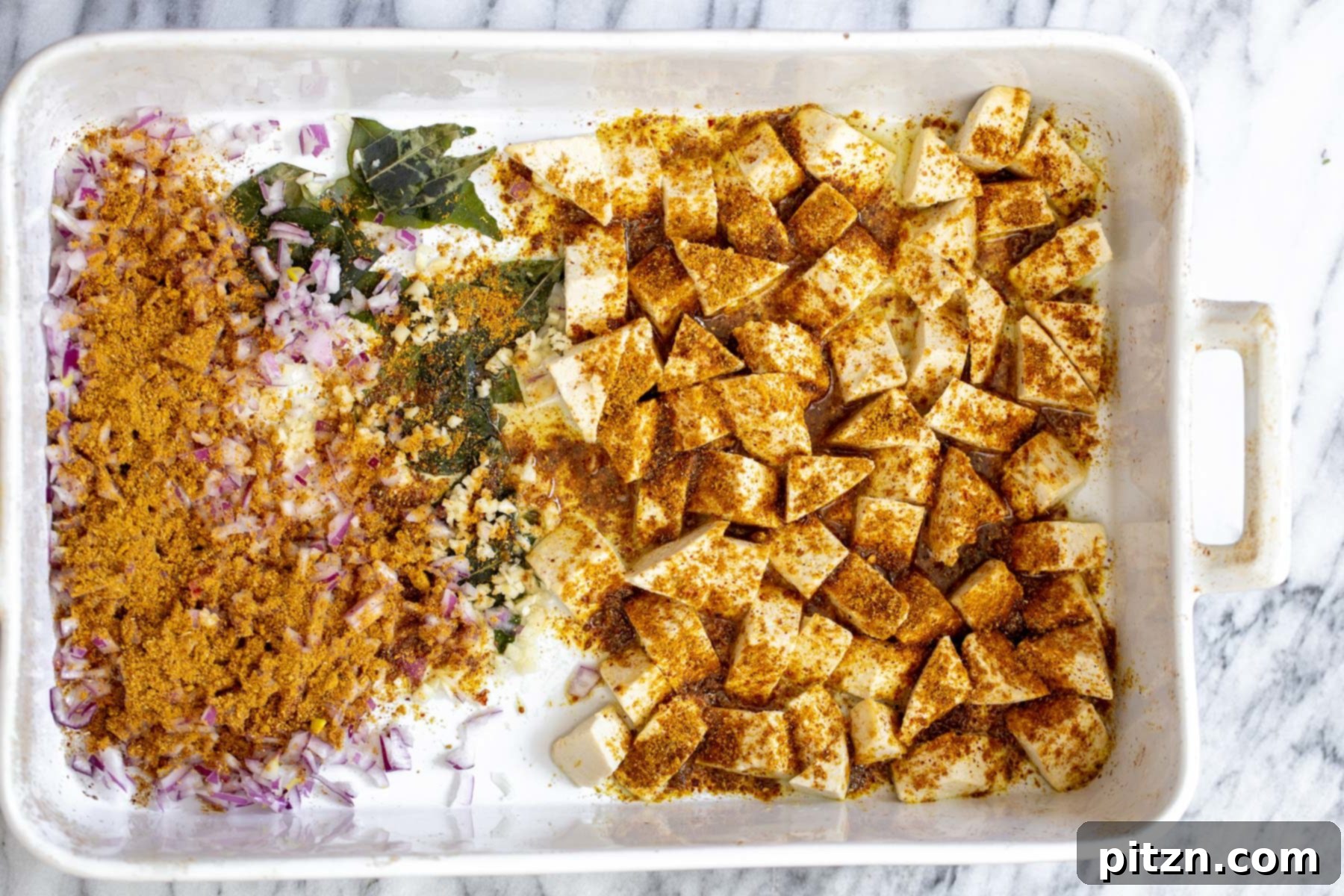 Tofu, onions, and curry leaves in a casserole dish, generously sprinkled with aromatic Madras curry powder before baking.