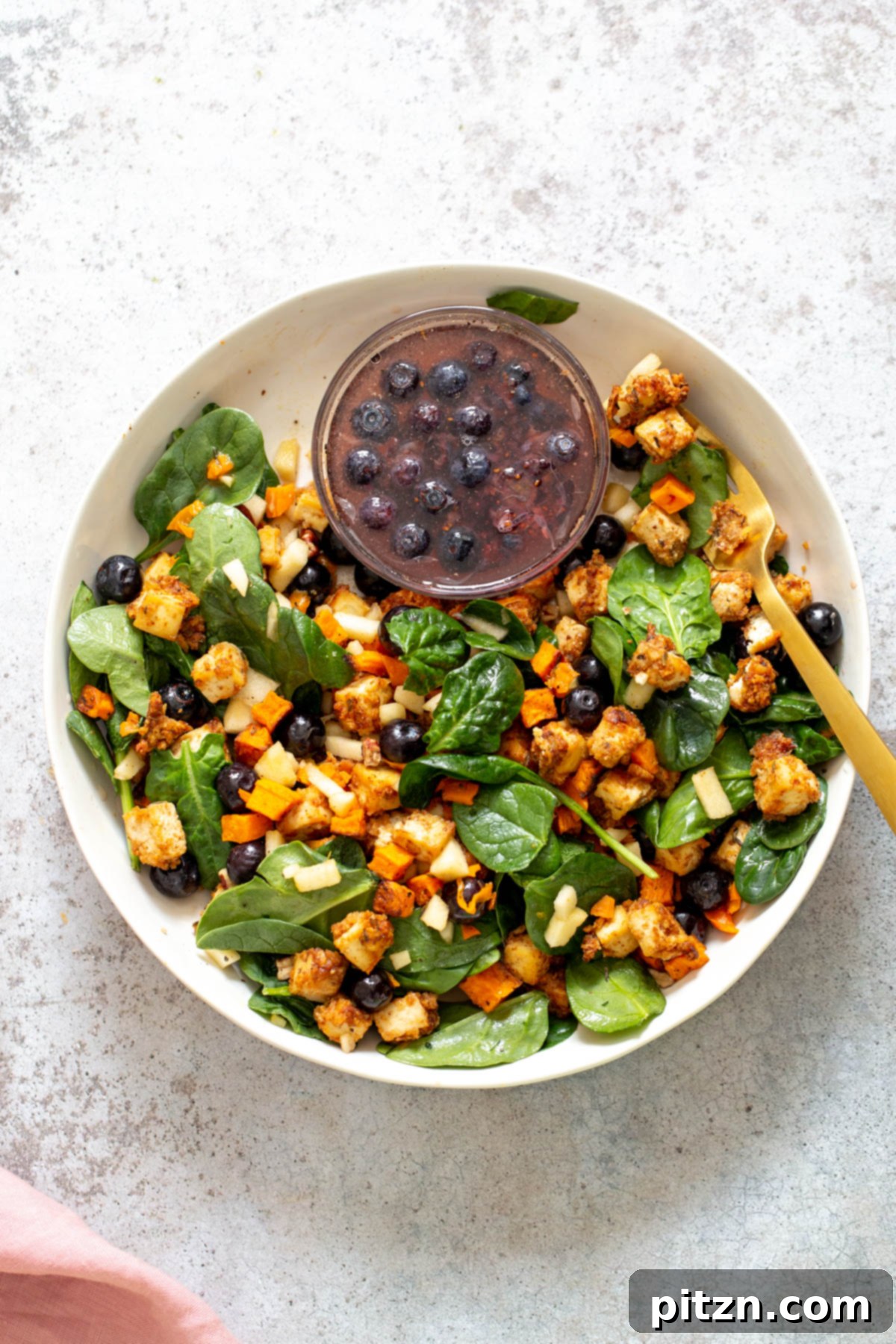 Sweet Potato Tofu Bowl A meticulously arranged vegan sweet potato tofu bowl featuring a base of baby spinach, topped with golden baked sweet potatoes, crispy tofu, and a side of maple blueberry dressing.