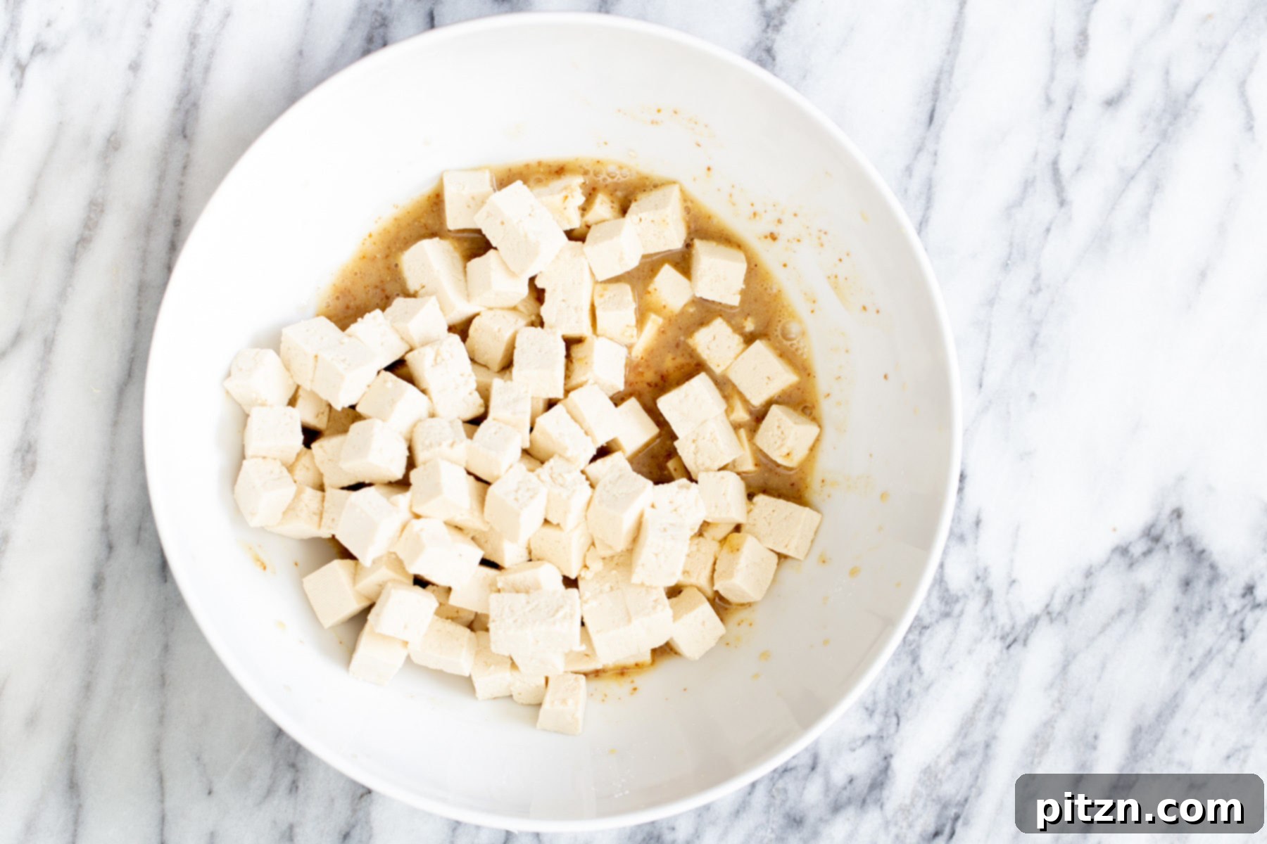 Spicy Maple Mustard Tofu and Sweet Potato Bowl 5 Cubed tofu being gently mixed with the rich maple mustard marinade in a white bowl, ensuring every piece is coated.