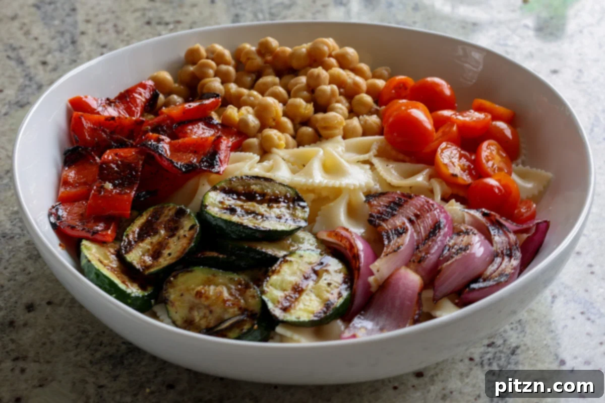 A colorful array of chickpeas, cooked pasta, and freshly grilled vegetables gathered in a large mixing bowl, ready for the dressing.