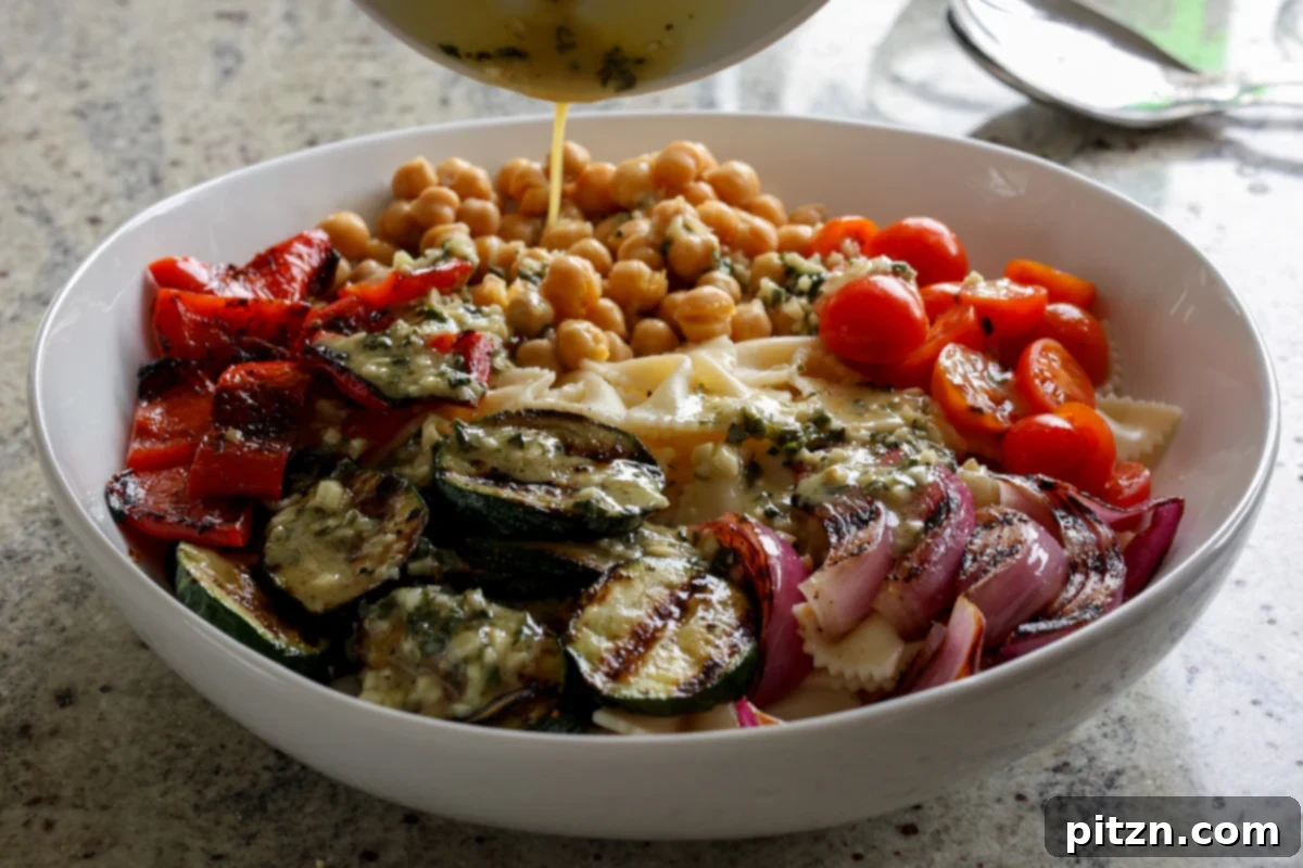The homemade Italian dressing being poured over the mixture of chickpeas, pasta, and grilled vegetables in a large mixing bowl.