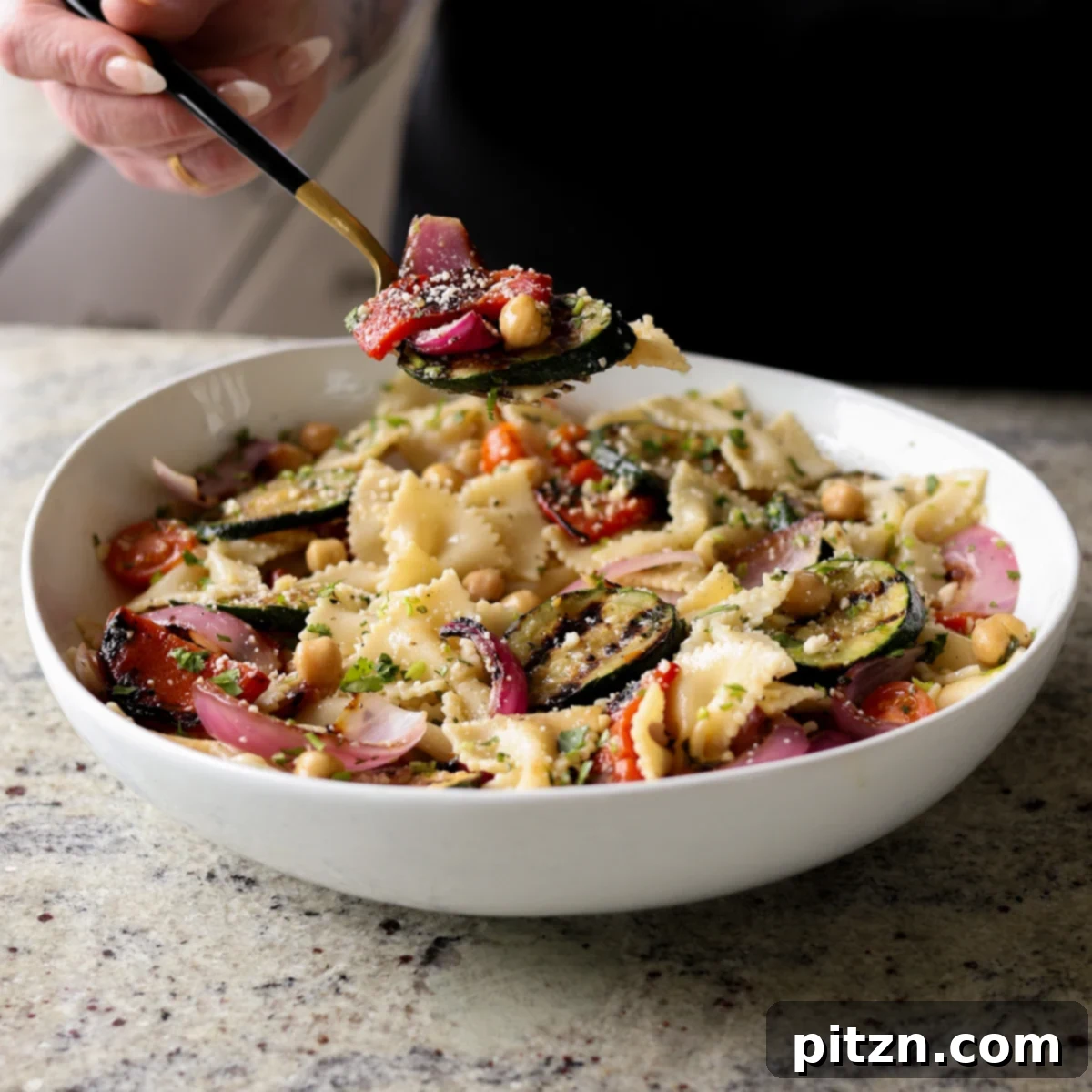 A spoon lifting a delightful bite of summer pasta salad, laden with grilled vegetables, pasta, and fresh herbs.
