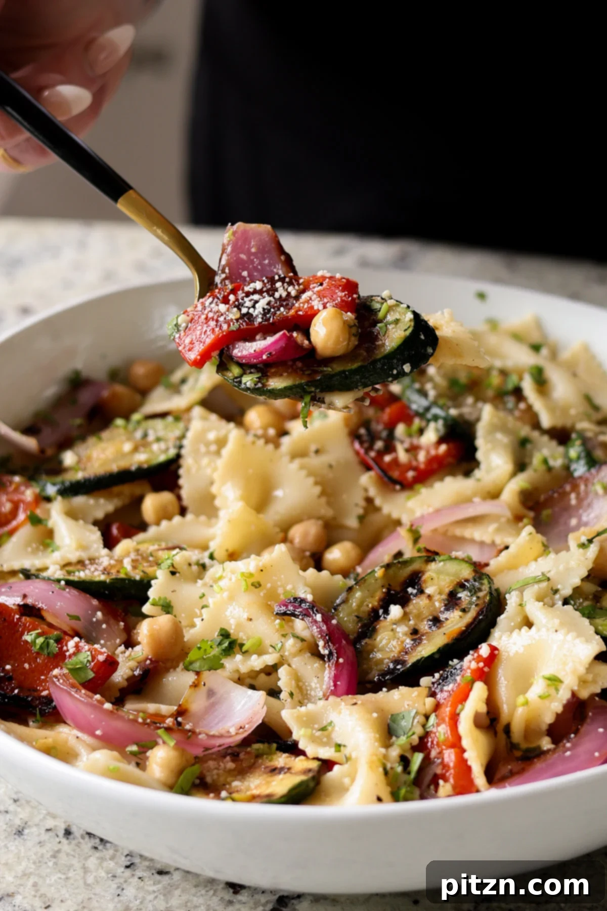 A spoon diving into a generous serving of summer pasta salad with grilled vegetables, showcasing the rich textures and vibrant colors.
