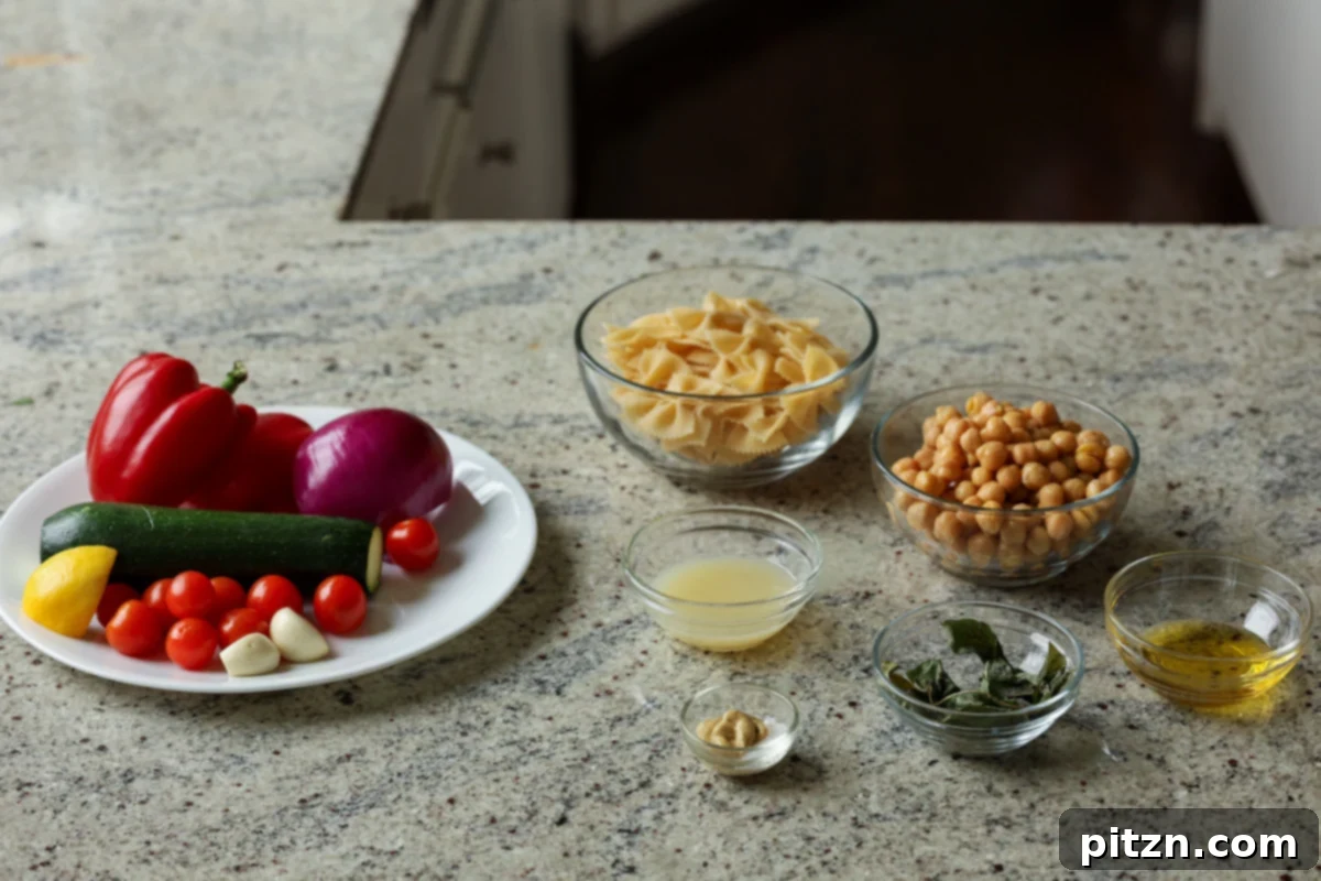 Various fresh ingredients for the pasta salad laid out on a kitchen counter, including raw pasta, fresh tomatoes, zucchini, bell peppers, onion, chickpeas, and herbs.