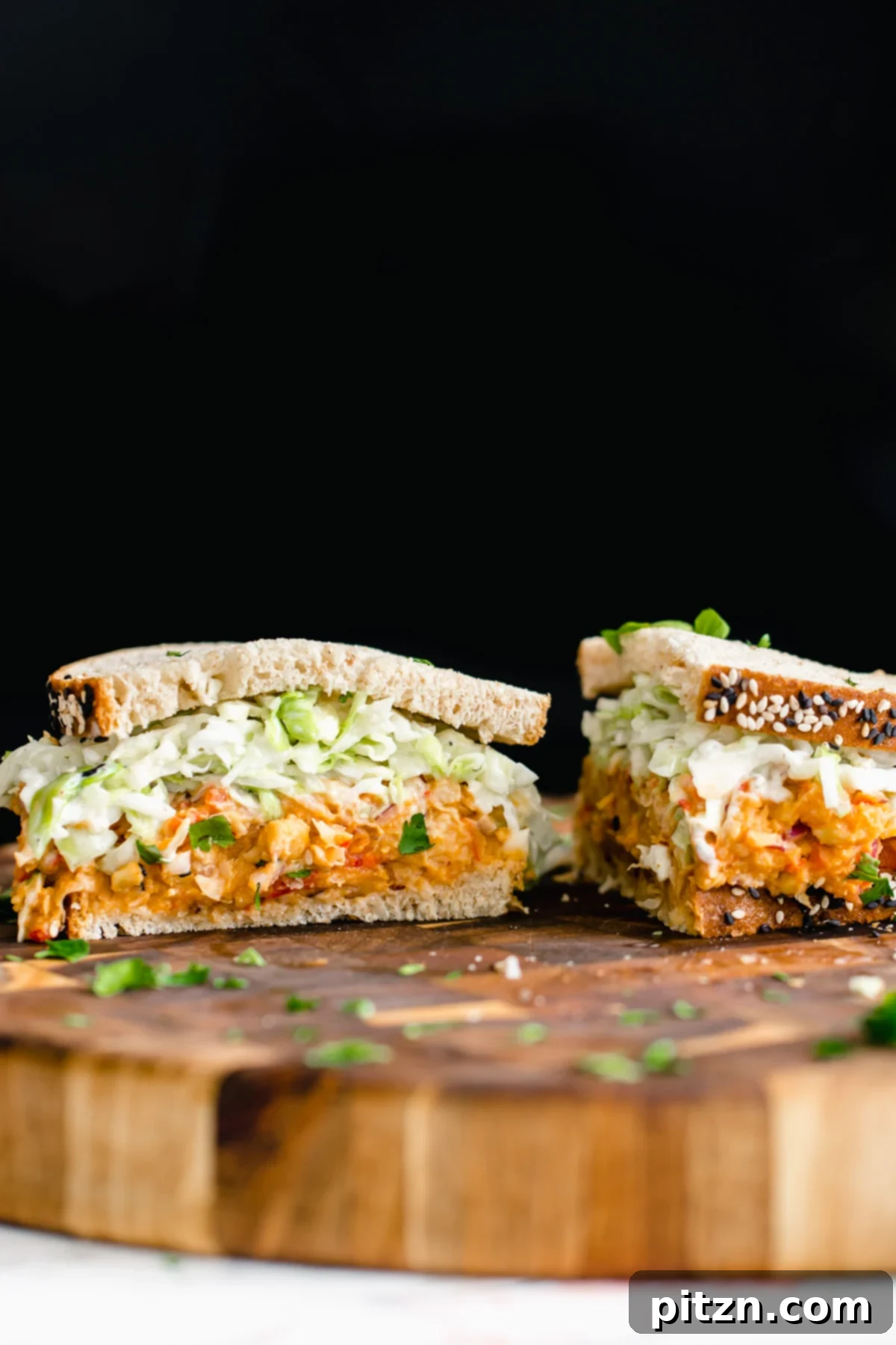 A beautifully halved Buffalo Chickpea Salad Sandwich with visible layers of spicy chickpea salad and crunchy slaw, resting on a wooden chopping board.
