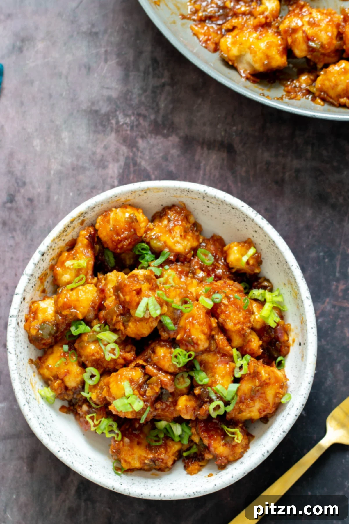 Golden Baked Cauliflower Manchurian 15 A generous serving of vegan Cauliflower Manchurian in a white bowl, garnished with green onions and a golden fork on the side